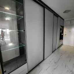 Nowoczesna szafa wnękowa z przesuwnymi, szarymi drzwiami i przeszkloną witryną z podświetlanymi półkami, w przedpokoju z marmurową podłogą. Widoczny fragment siedziska.