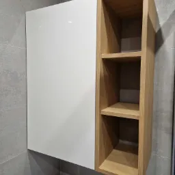 Asymetryczna szafka łazienkowa: biały, lakierowany front i otwarty regał z jasnego drewna dębowego. Nowoczesny design, szare płytki w tle, widoczny fragment umywalki.