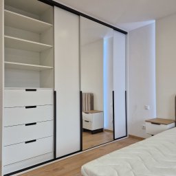 TOMEBEL - Biała szafa przesuwna z lustrem i czarnymi detalami w sypialni. Widoczne półki, szuflady i fragment łóżka. Minimalistyczny design, jasne wnętrze.