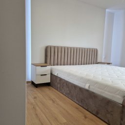 TOMEBEL - Beżowe łóżko tapicerowane z pionowymi przeszyciami wezgłowia i biała szafka nocna z drewnianym blatem na parkiecie w minimalistycznej sypialni.
