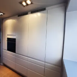 Nowoczesna kuchnia z zabudową w kolorze jasnego beżu, zintegrowana z piekarnikiem, widoczne drewniane panele podłogowe i oświetlenie sufitowe.