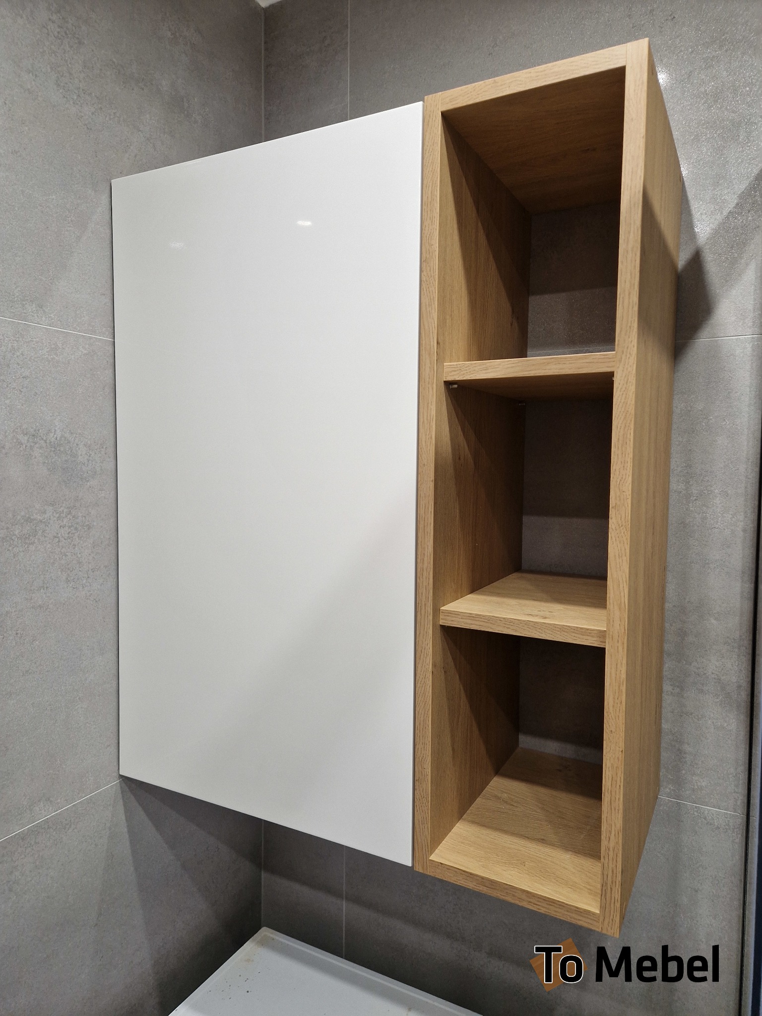 Asymetryczna szafka łazienkowa: biały, lakierowany front i otwarty regał z jasnego drewna dębowego. Nowoczesny design, szare płytki w tle, widoczny fragment umywalki.
