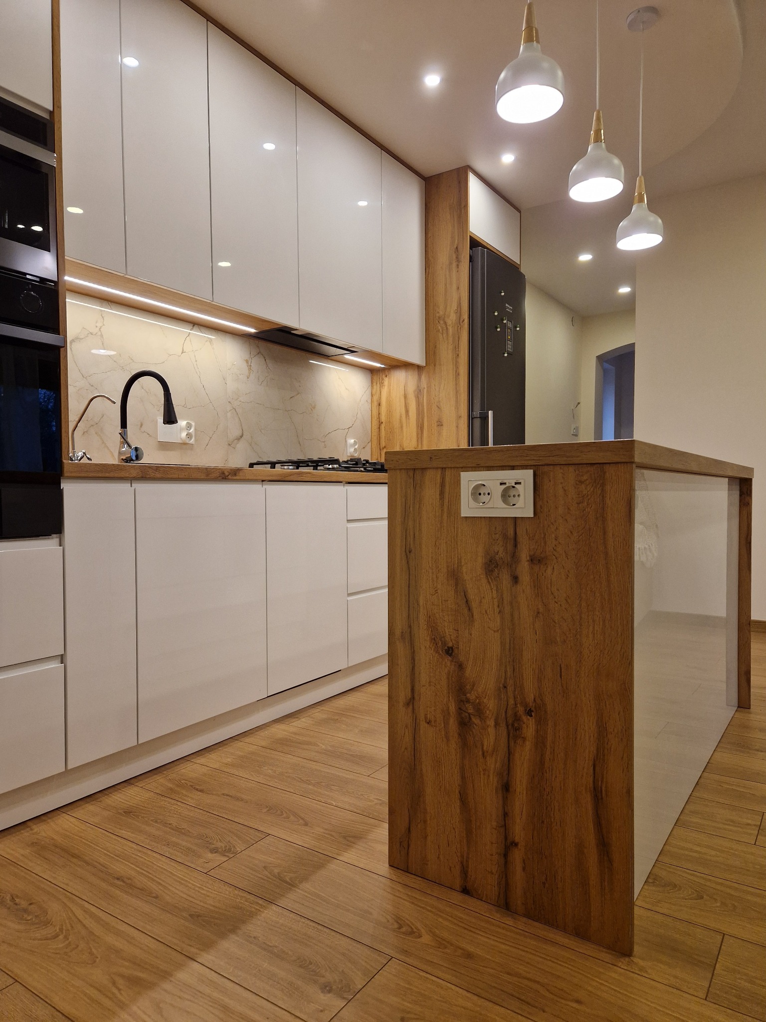 Nowoczesna kuchnia z wyspą: białe fronty szafek, drewniany blat i wyspa z gniazdkami. Oświetlenie punktowe i marmurowy backsplash dodają elegancji.