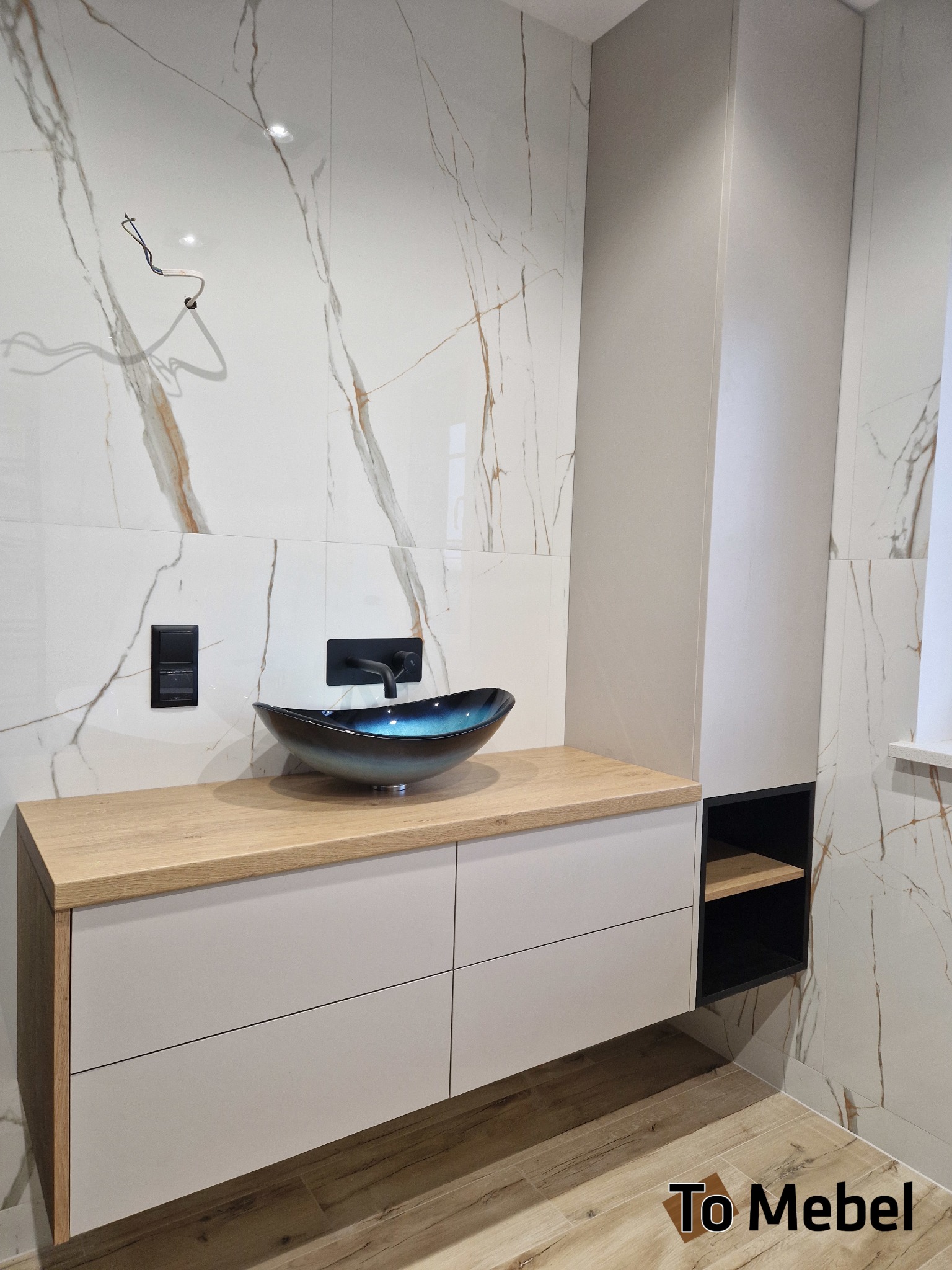 Nowoczesna szafka łazienkowa z drewnianym blatem i umywalką w kształcie misy na tle marmurowych płytek. Minimalistyczny design z czarnymi akcentami.