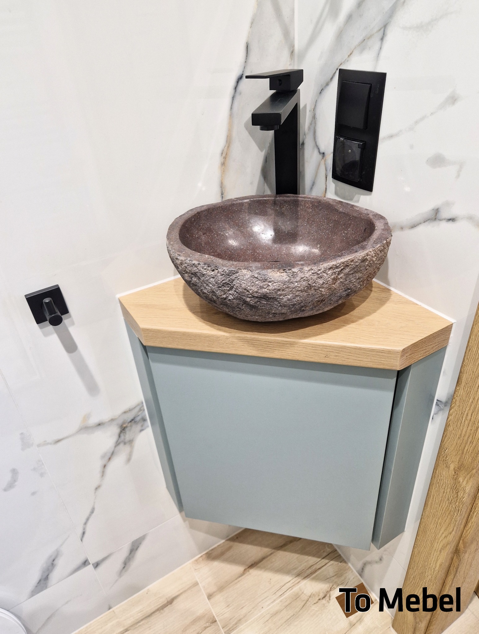 Narożna szafka łazienkowa z kamienną umywalką w stylu rustykalnym i czarną armaturą na tle marmurowej ściany. Minimalistyczny design z akcentem naturalnych materiałów.