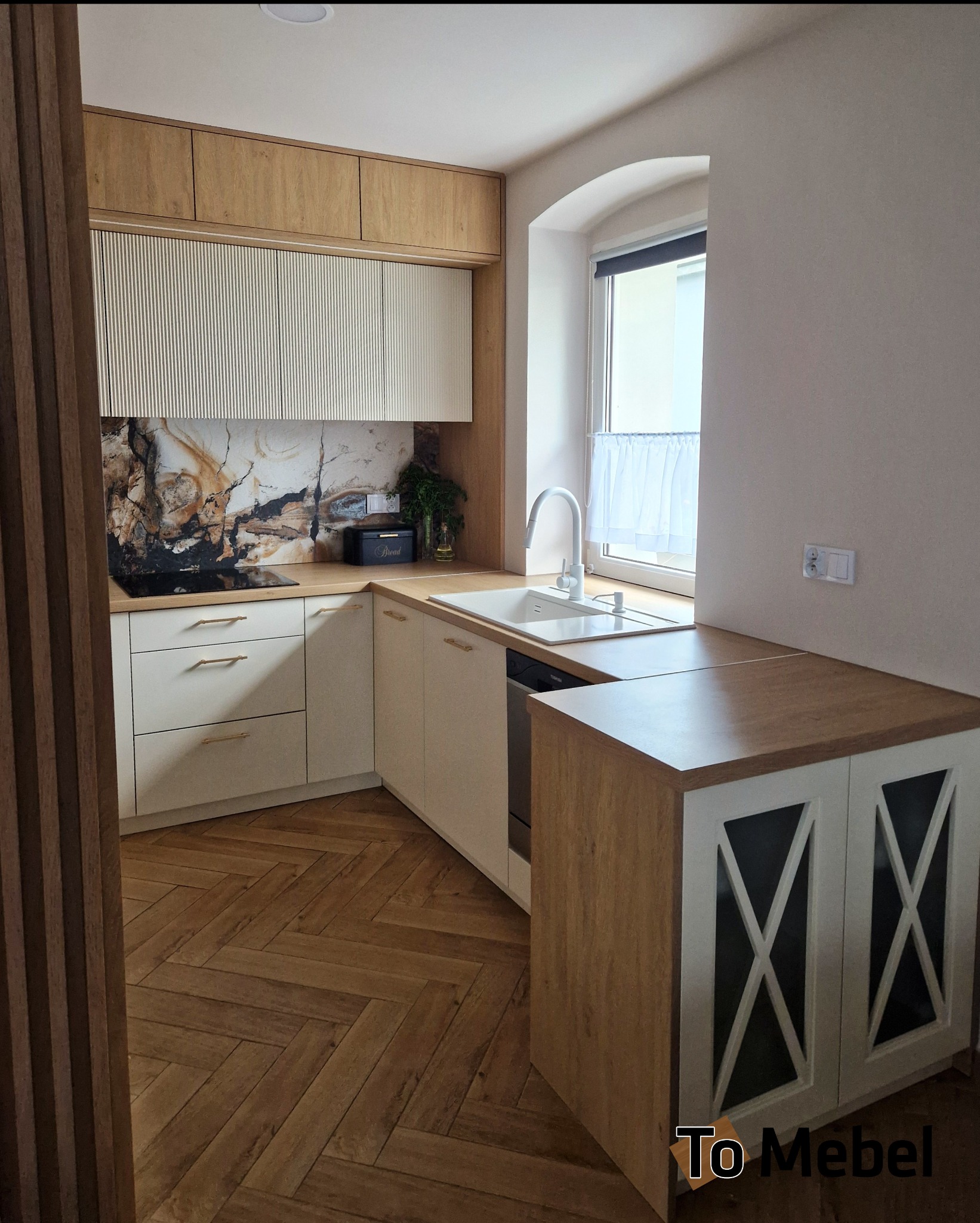 Jasna kuchnia z drewnianymi akcentami: frezowane fronty szafek górnych, marmurowy backsplash, parkiet w jodełkę. Ujęcie z perspektywy wejścia do pomieszczenia.