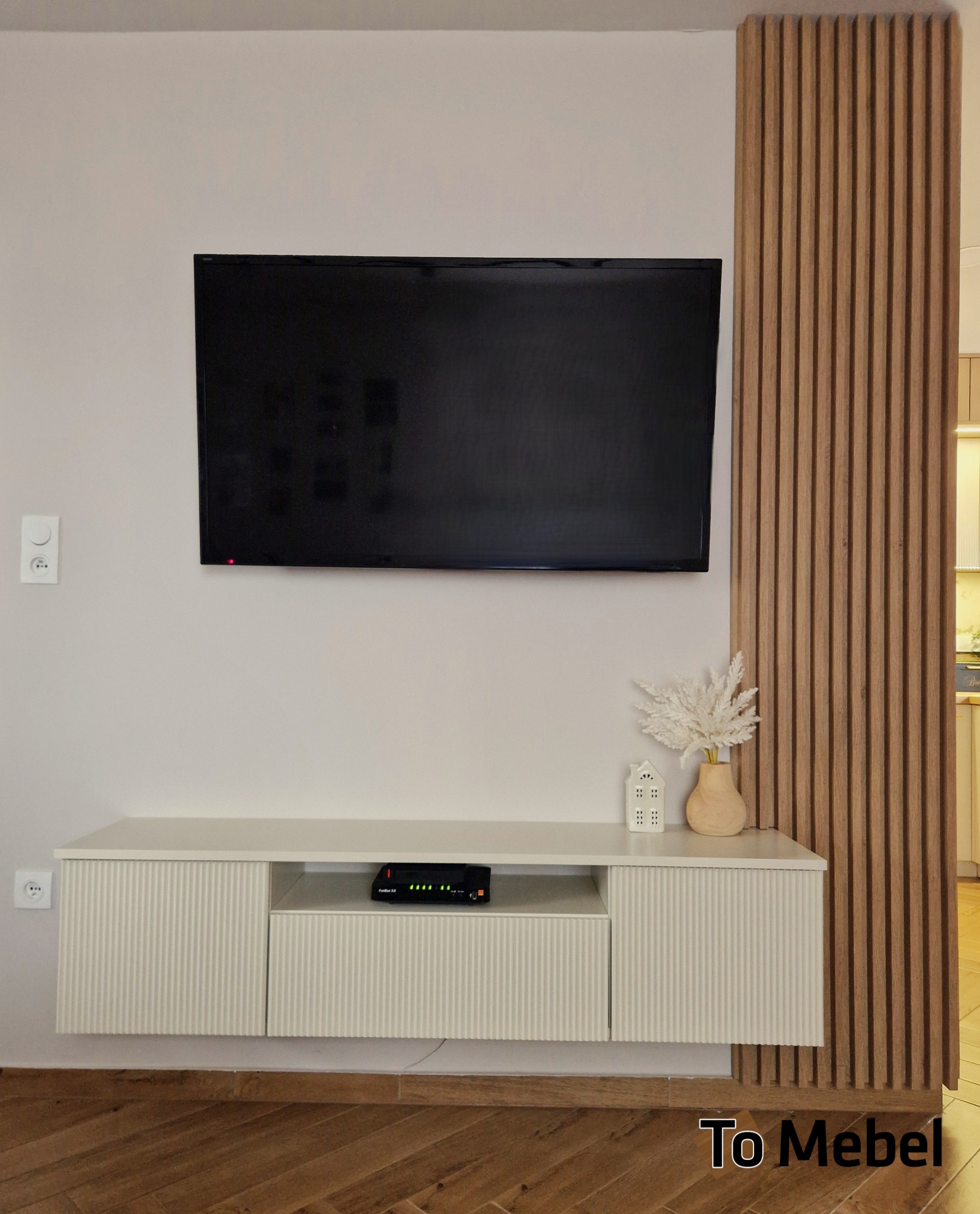 Biała szafka RTV z ryflowanymi frontami, wisząca pod telewizorem, z dekoracjami i drewnianą ścianką działową. Nowoczesny design, ciepłe oświetlenie.