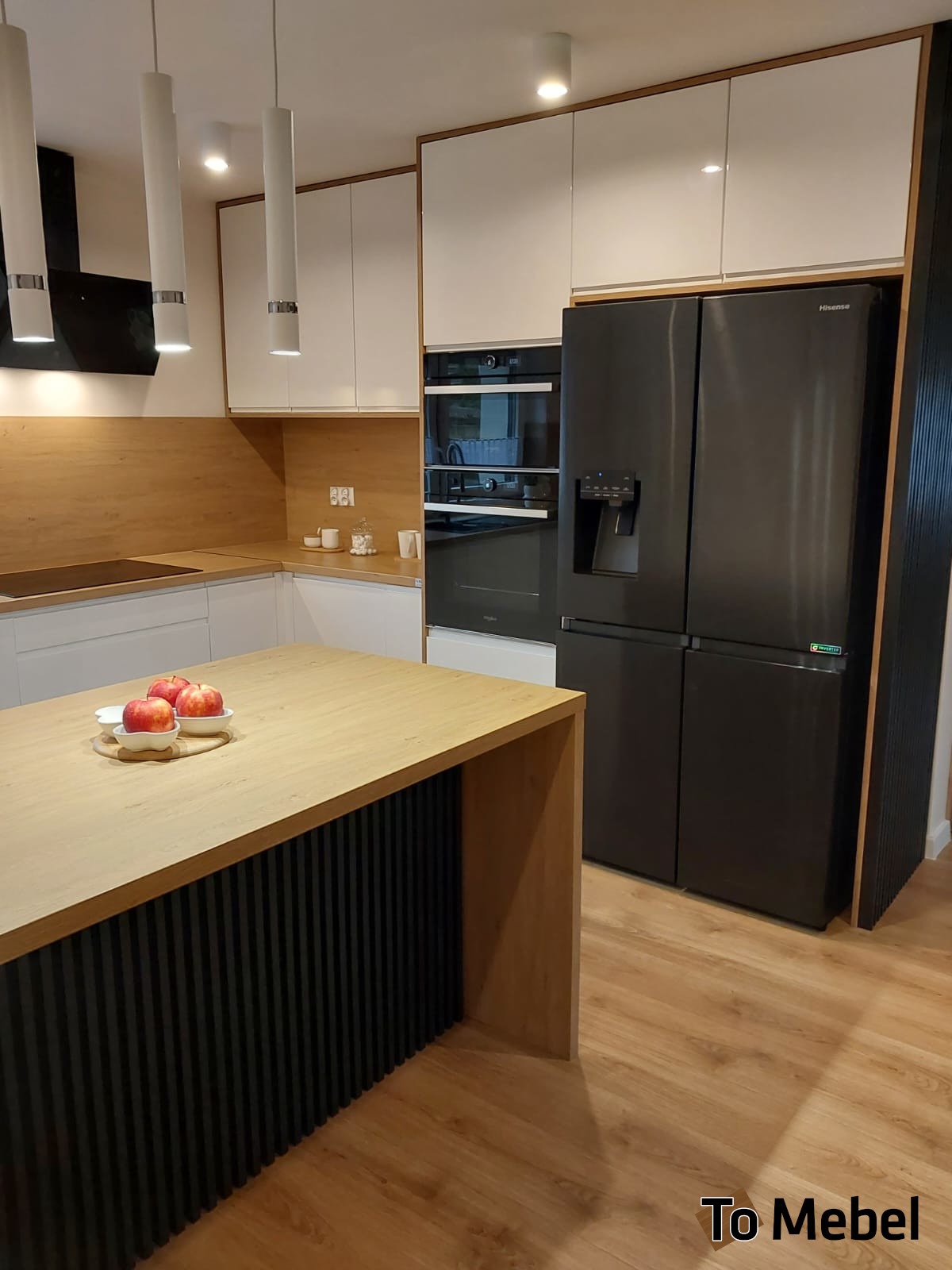 Nowoczesna kuchnia z wyspą: białe szafki, drewniany blat, czarny sprzęt AGD. Na wyspie jabłka w miseczkach. Minimalistyczne lampy wiszące.