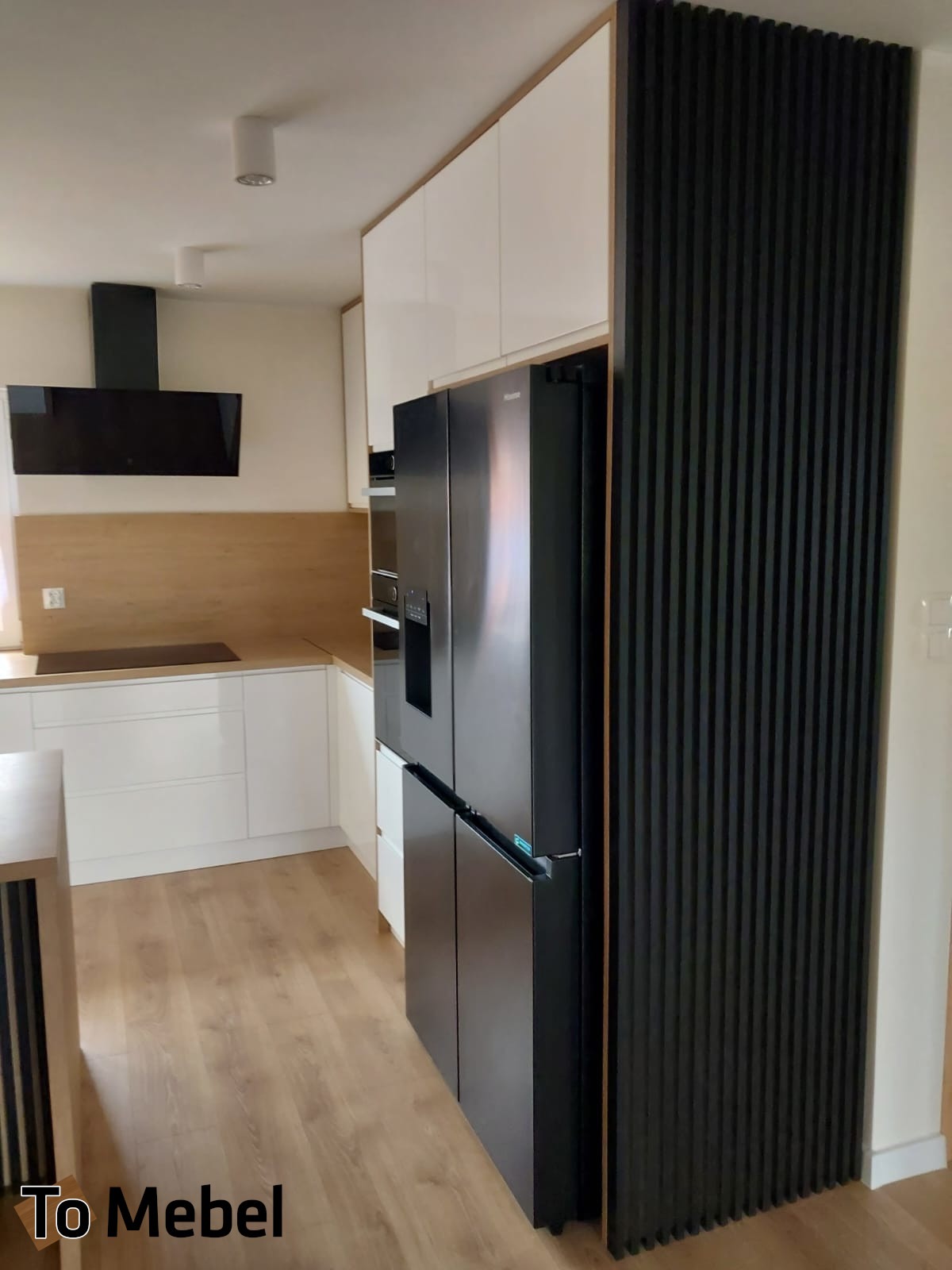 Nowoczesna kuchnia z białymi szafkami, drewnianym blatem i czarną lodówką obudowaną lamelami. Widoczny fragment wyspy kuchennej i okap.