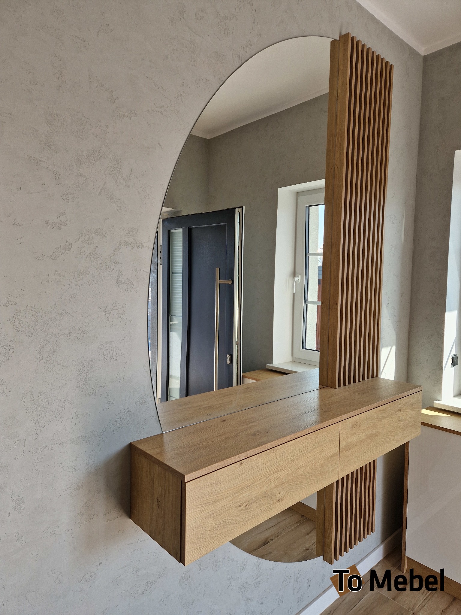 Nowoczesna konsola z lustrem w kształcie półkola i drewnianymi lamelami, zawieszona na szarej ścianie. Widoczne drzwi i okno w tle. Jasne drewno, minimalistyczny design.