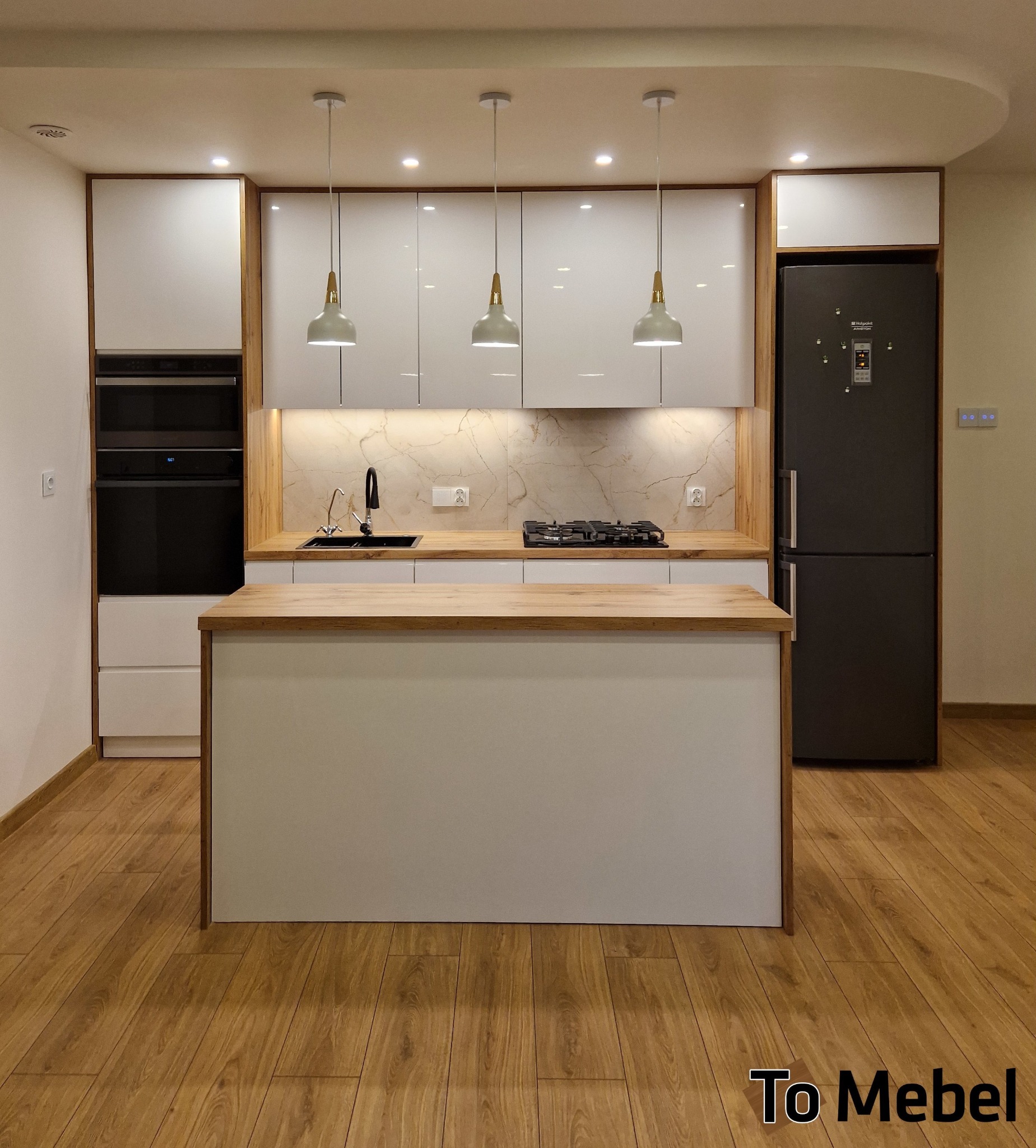 Nowoczesna kuchnia na wymiar z wyspą, białe fronty szafek, drewniany blat i okładzina ściany, czarna lodówka i piekarnik w zabudowie. Oświetlenie punktowe i wiszące.