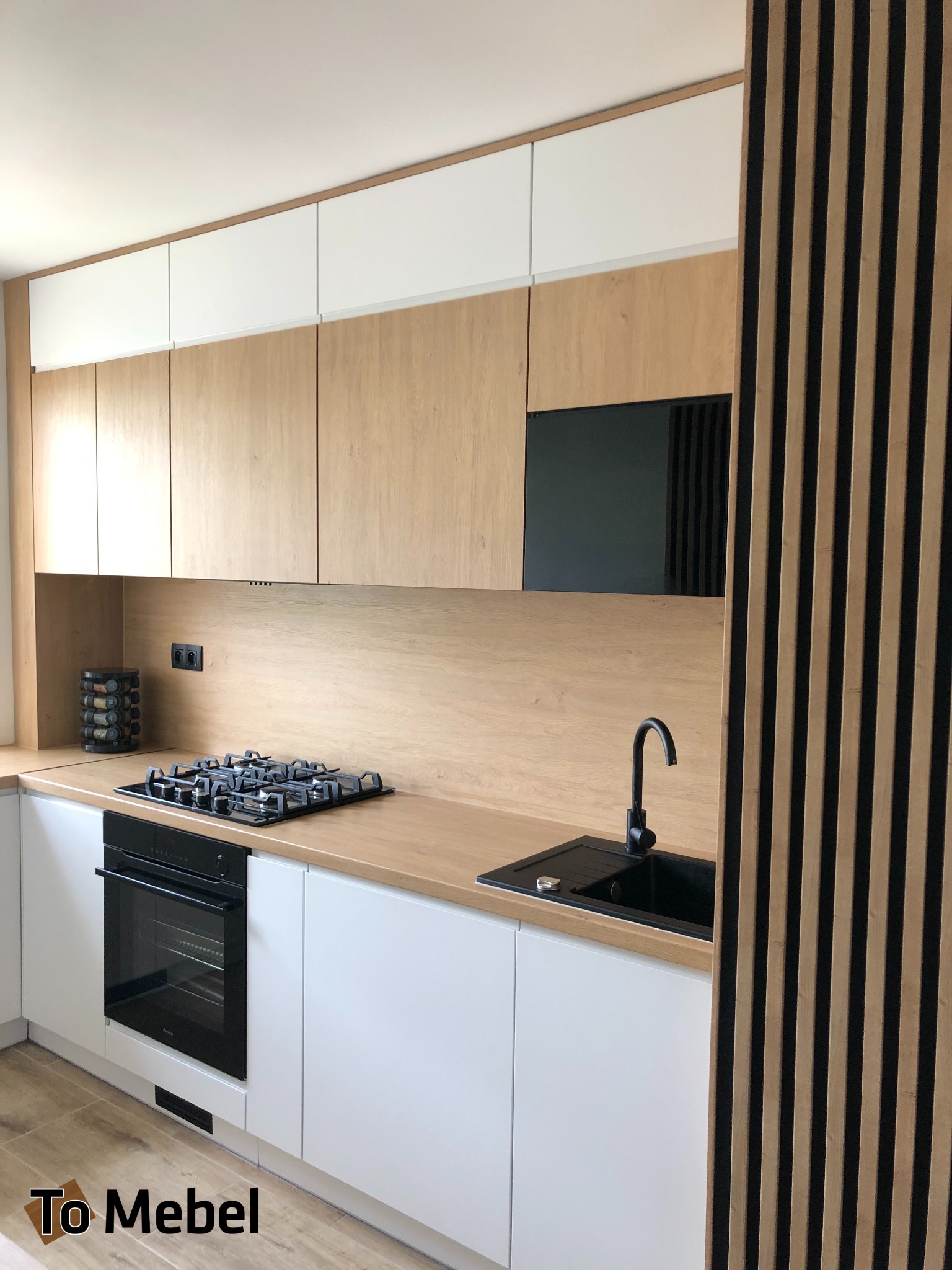 Jasna kuchnia z drewnianymi frontami i białymi szafkami, czarny zlew i płyta gazowa. Dekoracyjne lamele z drewna na ścianie. Nowoczesny design.