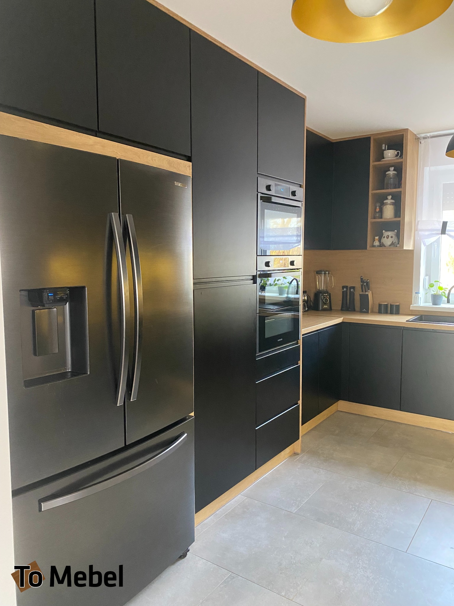 Nowoczesna kuchnia z czarnymi frontami i drewnianymi akcentami. Widoczna zabudowa z lodówką side-by-side, piekarnikiem i mikrofalówką w słupku oraz szafki wiszące.