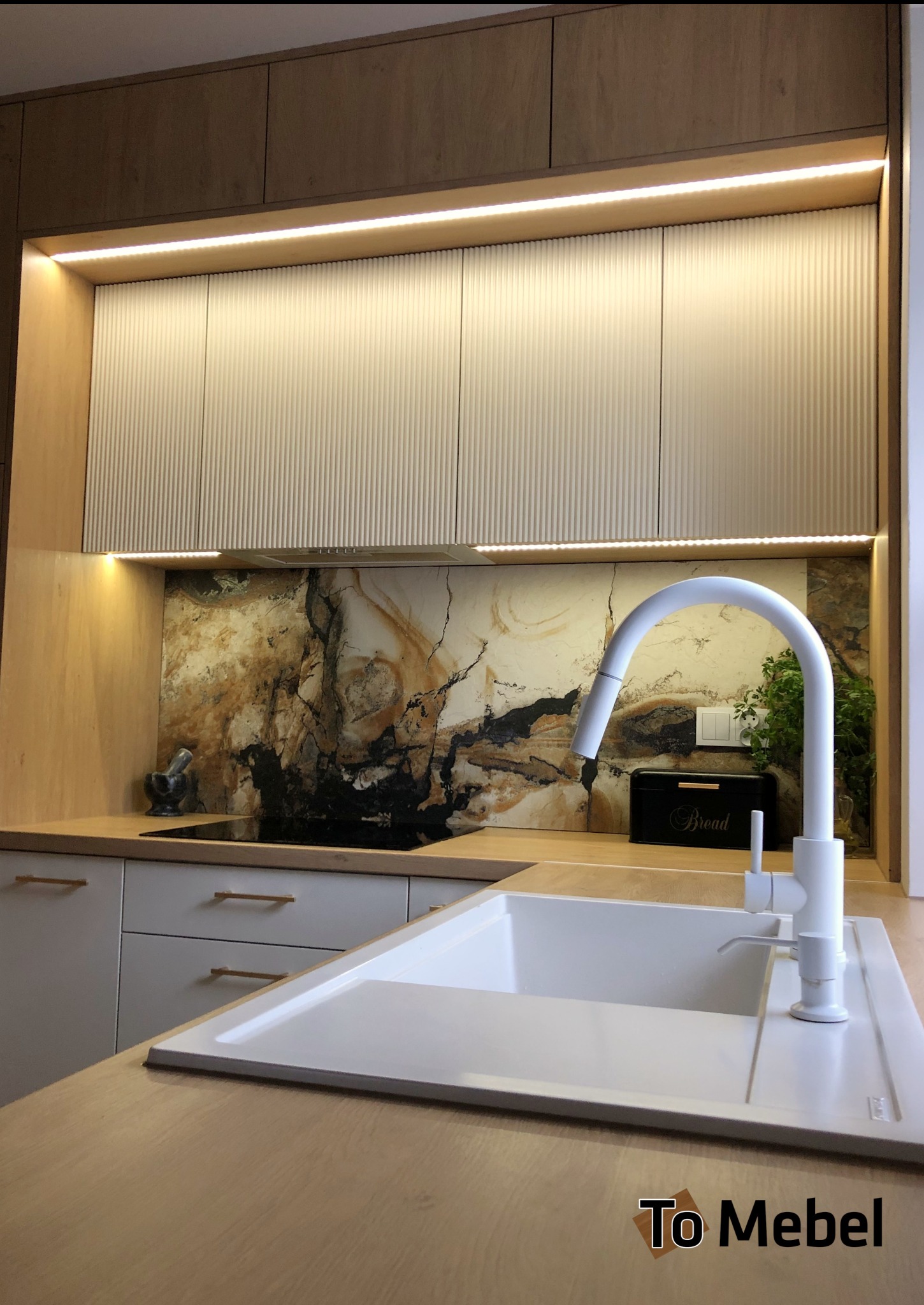 Nowoczesna kuchnia z frezowanymi frontami szafek górnych, podświetleniem LED i blatem imitującym drewno. Uwagę zwraca marmurowy panel nad blatem i biała armatura.