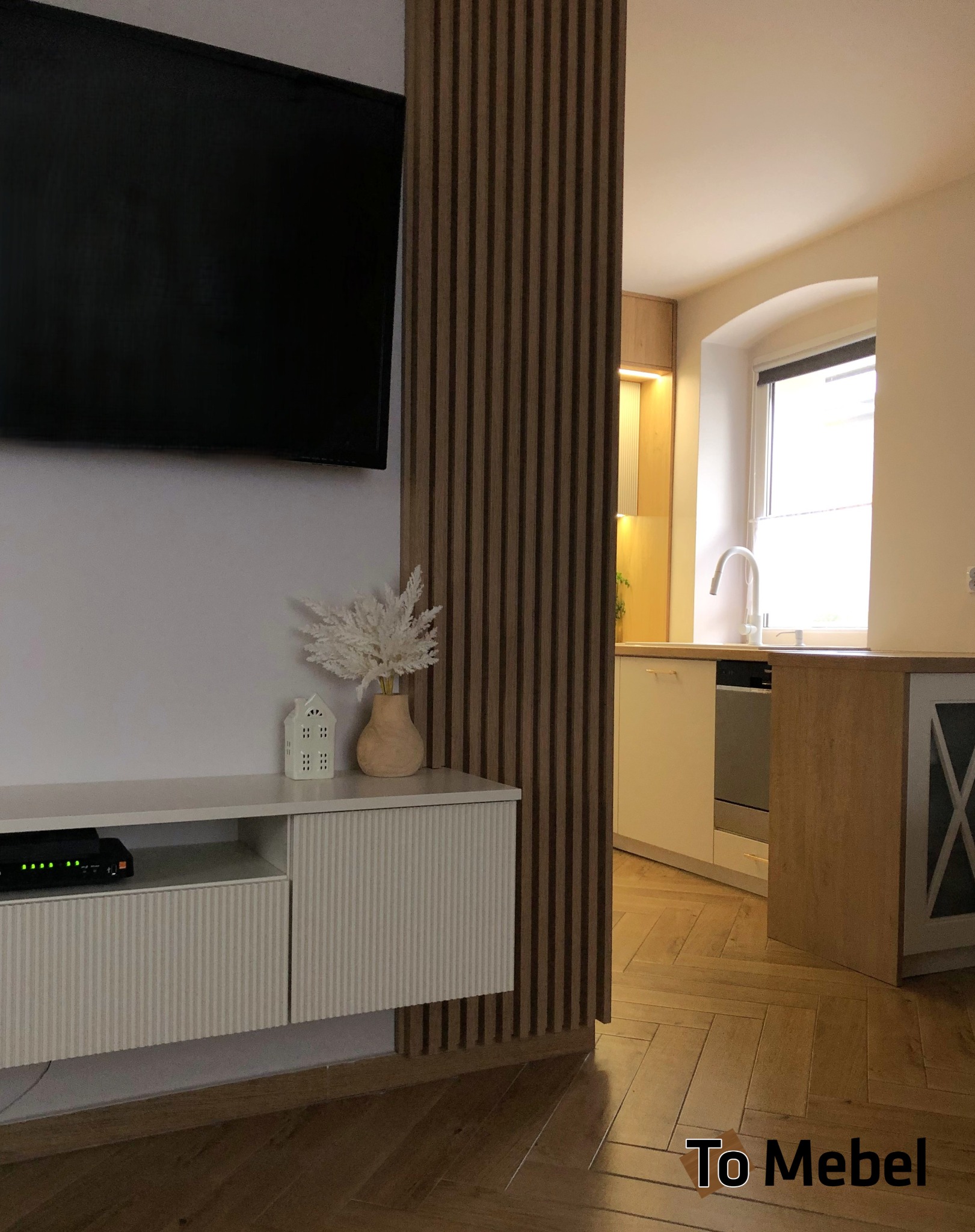 Nowoczesny salon z telewizorem i szafką RTV, oddzielony od kuchni drewnianą ścianką lamelową. W tle widoczna zabudowa kuchenna z oknem.