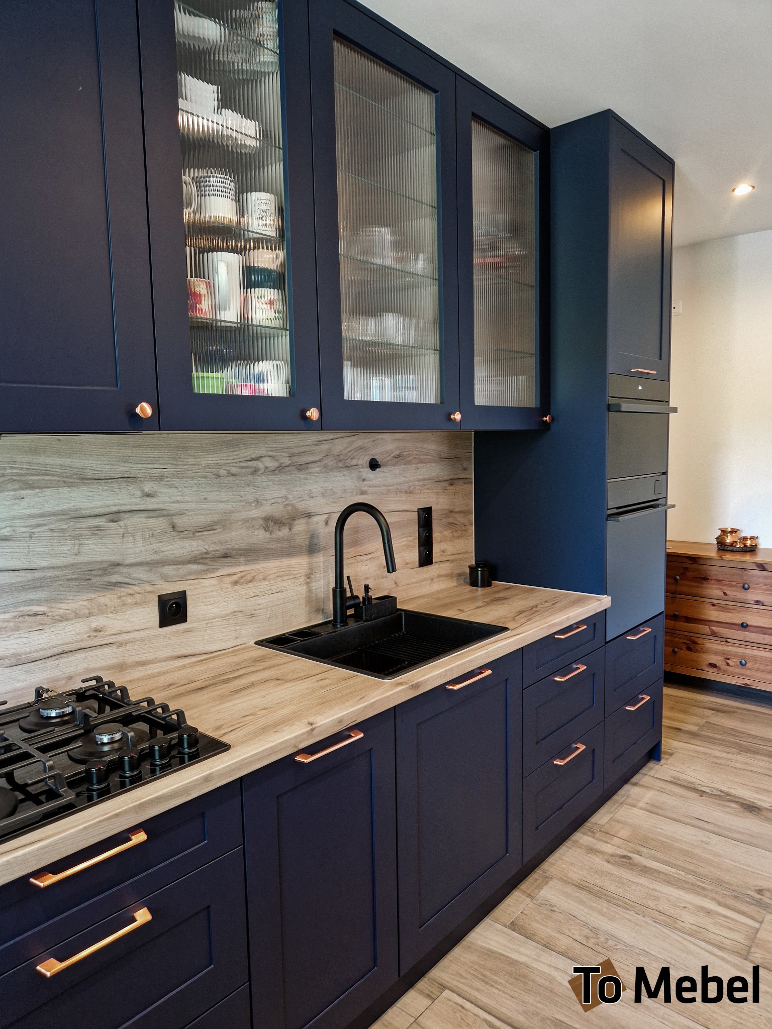 Granatowa zabudowa kuchenna z ryflowanymi szybami, drewnianym blatem i miedzianymi uchwytami. Czarny zlew i płyta gazowa. Ujęcie z perspektywy.