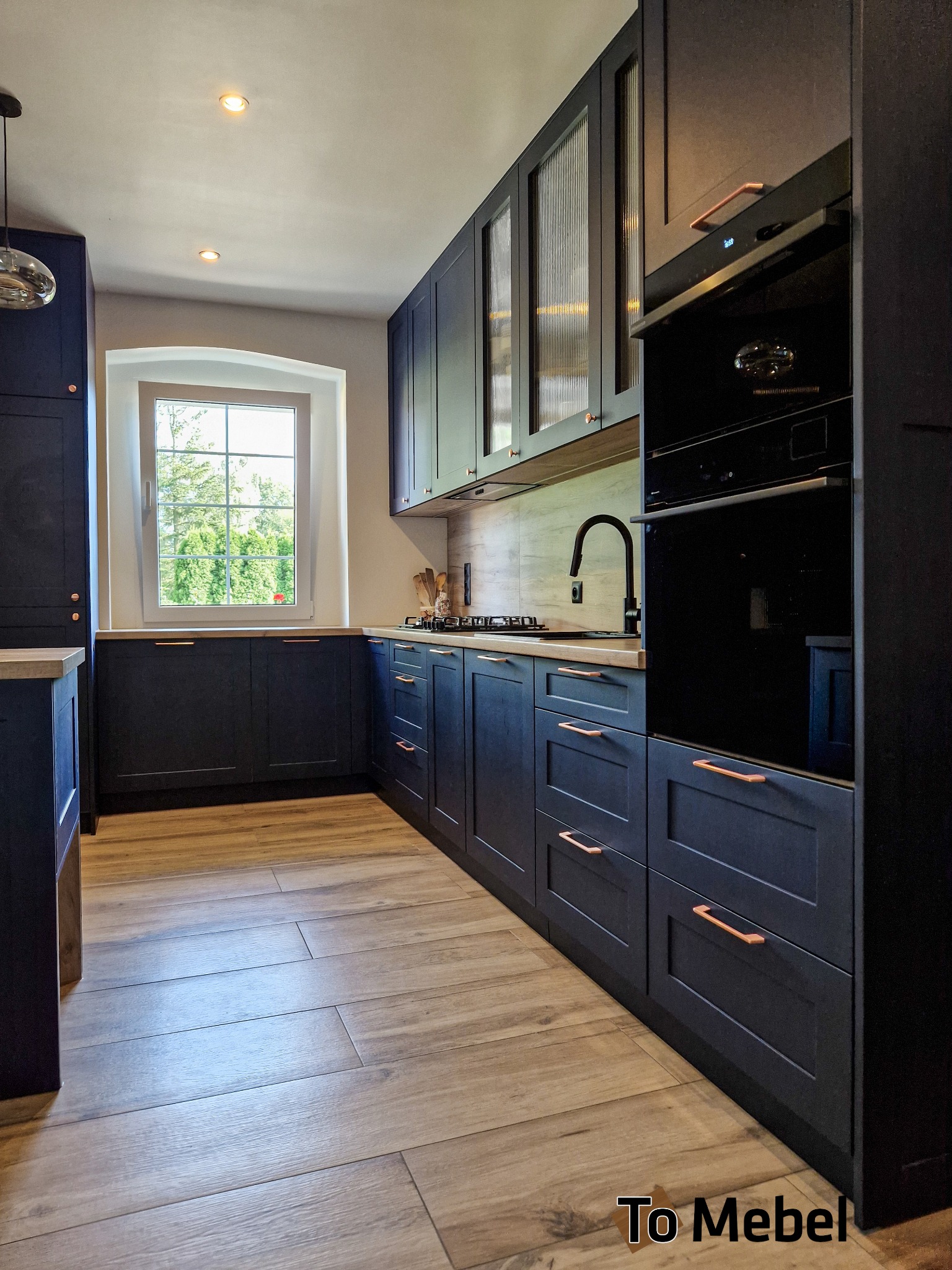 Ciemna kuchnia z drewnianymi blatami i miedzianymi uchwytami, oświetlona światłem dziennym z okna. Widoczne szklane fronty szafek i nowoczesny piekarnik.