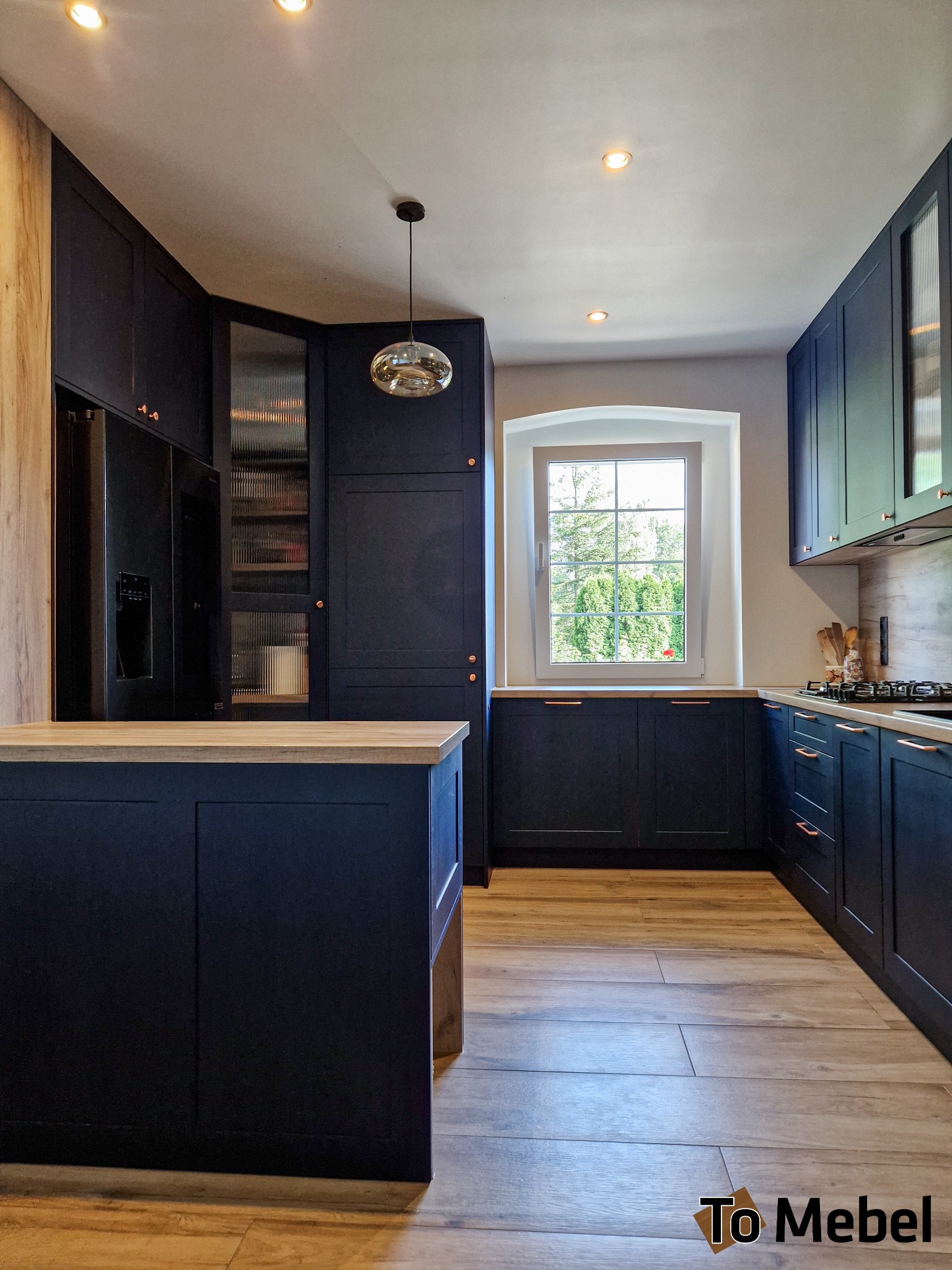 Granatowa kuchnia na wymiar z drewnianym blatem i wyspą, oświetlona lampą wiszącą. Widok na okno z zielenią. Nowoczesny design z akcentami miedzianymi.