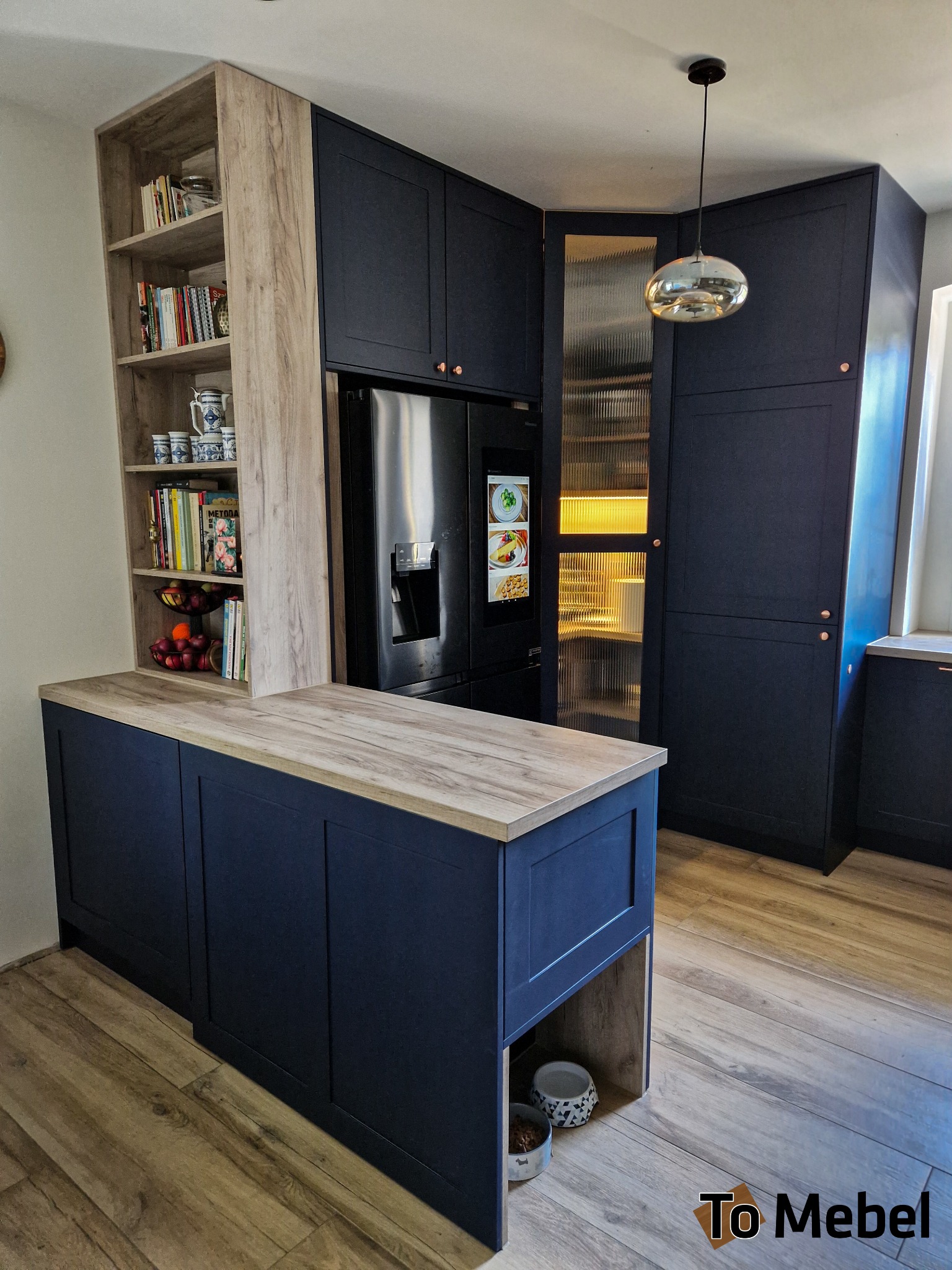 Granatowa zabudowa kuchenna z drewnianym blatem i regałem, oświetlona lampą wiszącą. Widoczna lodówka z wyświetlaczem i szafka z ryflowanym szkłem.