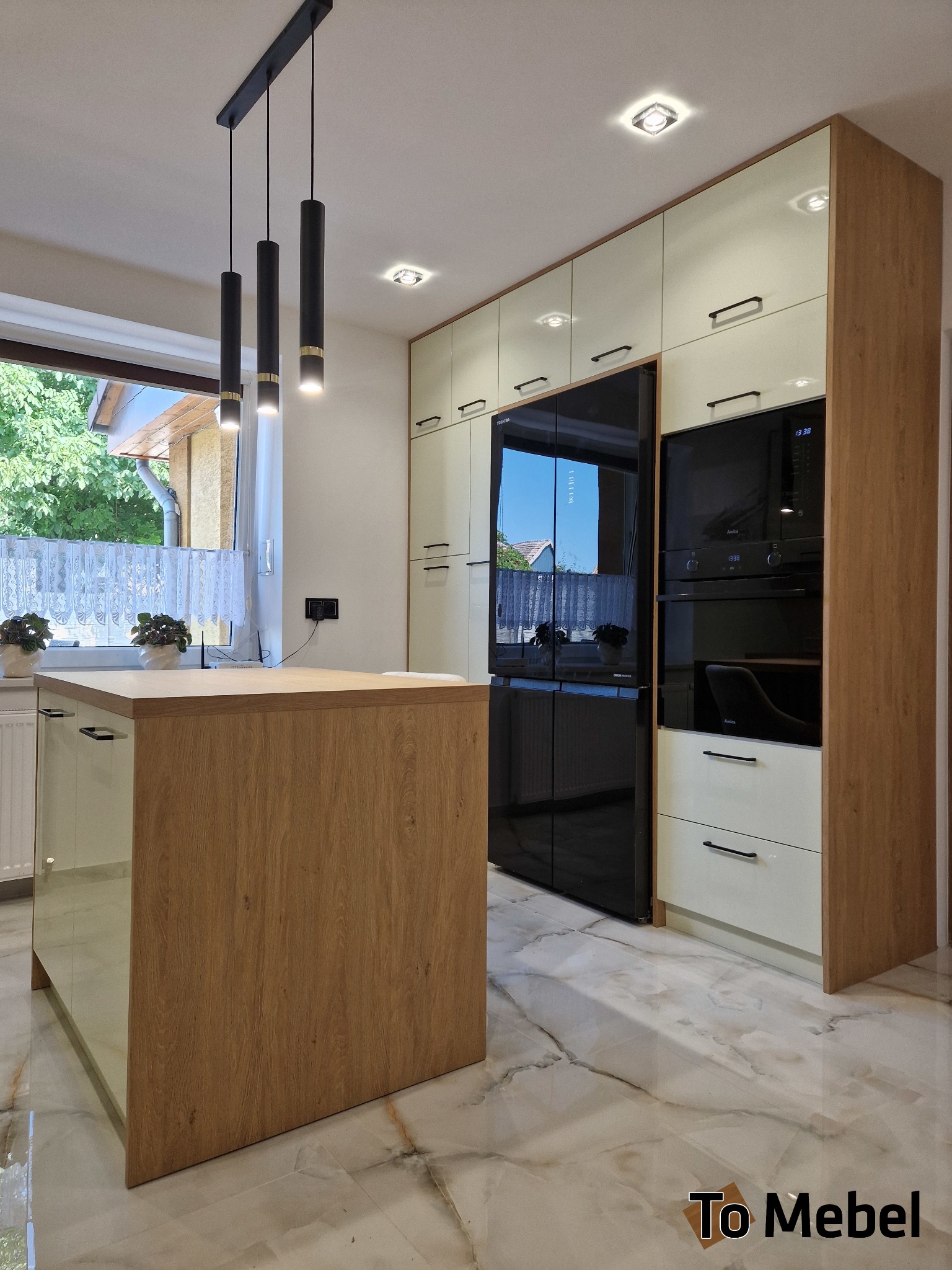 Nowoczesna kuchnia z wyspą: kremowe szafki, drewniane akcenty, czarny sprzęt AGD i marmurowa podłoga. Minimalistyczne lampy wiszące nad wyspą.