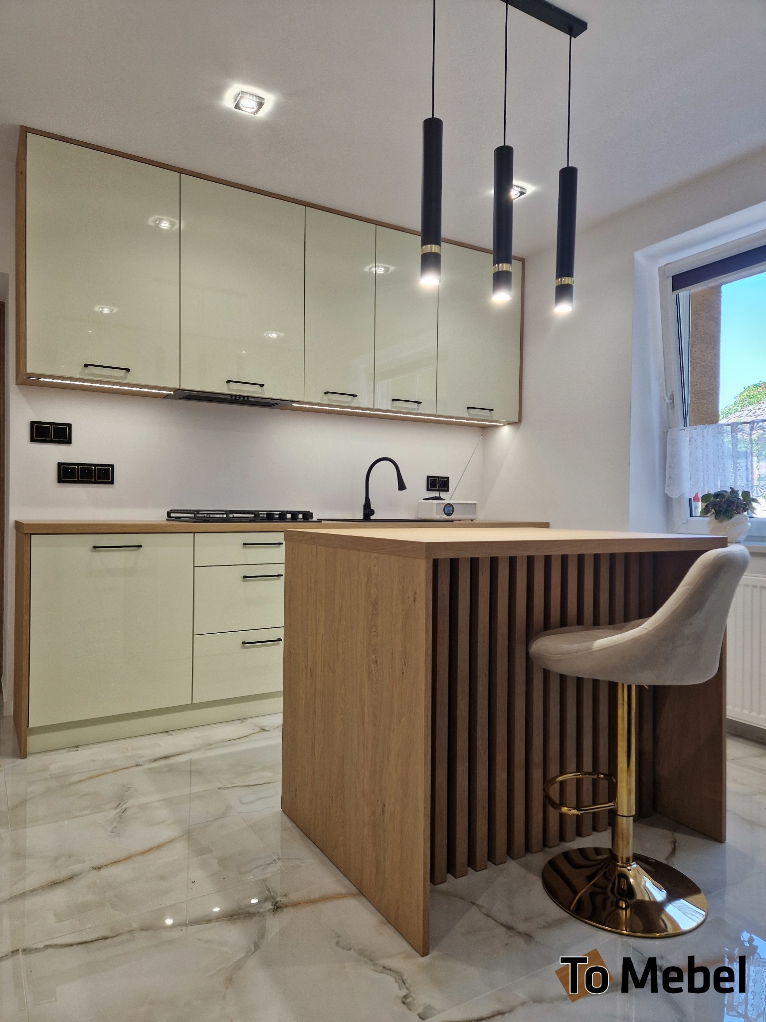 Jasna kuchnia z wyspą i hokerem na złotym stelażu. Górne szafki z oświetleniem LED, czarne, wiszące lampy. Marmurowe płytki podłogowe z wyraźnym wzorem.