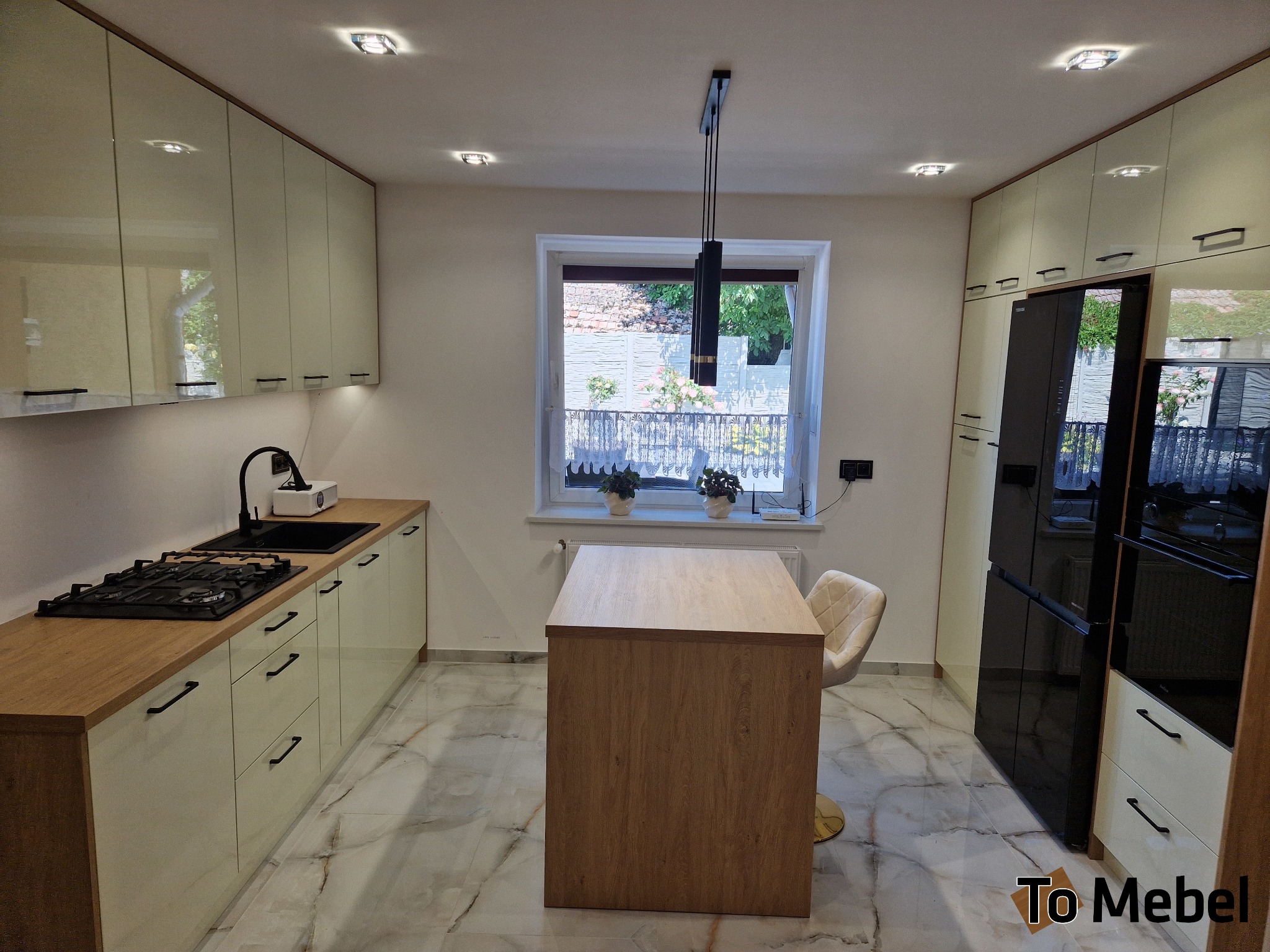 Jasna kuchnia z wyspą i czarnymi akcentami: bateria, zlew, płyta gazowa. Białe fronty szafek, drewniany blat i wyspa. Okno z widokiem na zieleń. Nowoczesny design.