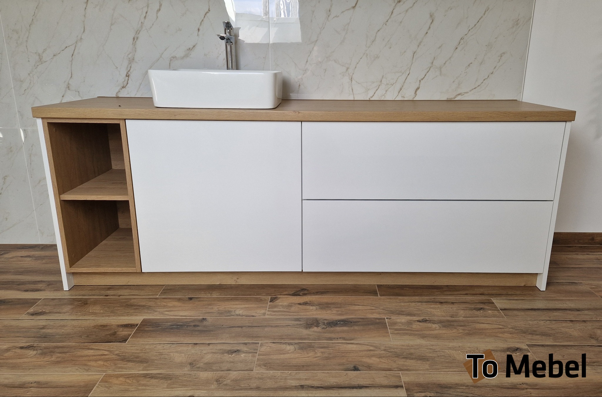 Nowoczesna szafka łazienkowa z białymi frontami i drewnianym blatem, na którym stoi biała umywalka. Po lewej stronie otwarte półki. Marmurowe płytki w tle.