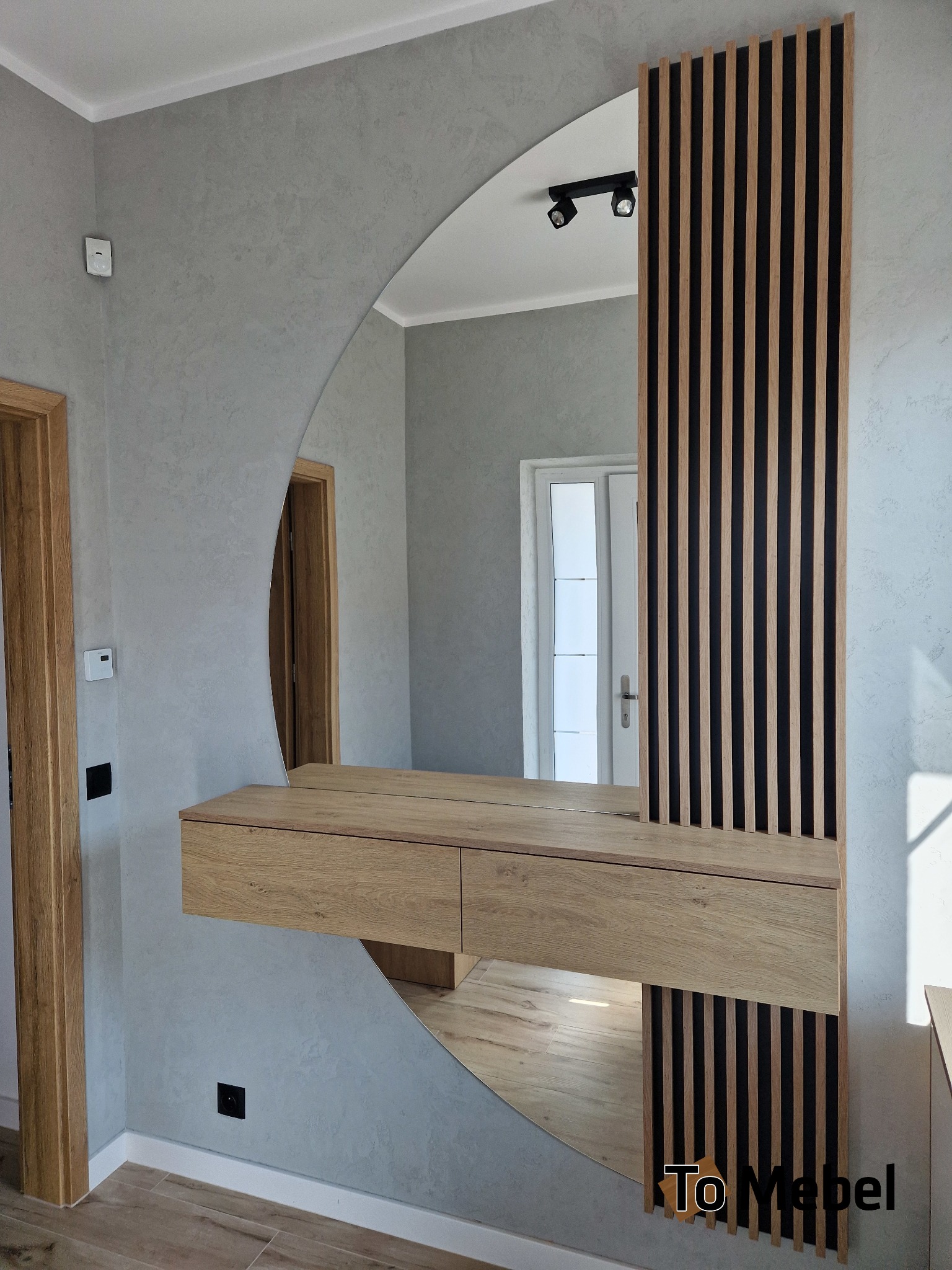 Nowoczesna szafka z lustrem w kształcie półkola i dekoracyjnymi lamelami w odcieniach drewna i czerni, zamontowana na szarej ścianie w przedpokoju.