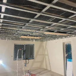 Montaż metalowej konstrukcji sufitu podwieszanego z izolacją akustyczną z wełny mineralnej, widoczna drabina aluminiowa, lampa halogenowa i narzędzia na podłodze w pomieszczeniu w trakcie remontu.