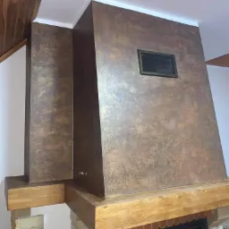 Ozdobna ściana kominkowa z efektem patyny w odcieniach brązu i złota, wykończona drewnianymi belkami i kamienną podstawą, widoczna kratka wentylacyjna.