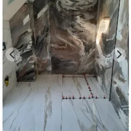 Układanie płytek podłogowych w łazience z użyciem systemu poziomowania, widoczne płytki imitujące marmur na podłodze i ścianach z wzorem przypominającym fale, w tle otwór na półkę w ścianie.