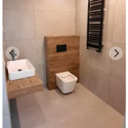 Nowoczesna łazienka z beżowymi płytkami, wiszącą umywalką na drewnianym blacie, podwieszaną toaletą z drewnianą obudową spłuczki i czarnym grzejnikiem drabinkowym.