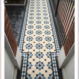 Wzorzysta, kremowo-niebiesko-czarna podłoga z płytek w stylu wiktoriańskim, ułożona na wąskim korytarzu w Choszcznie, widok z góry, z elementami muru z czerwonej cegły po bokach.