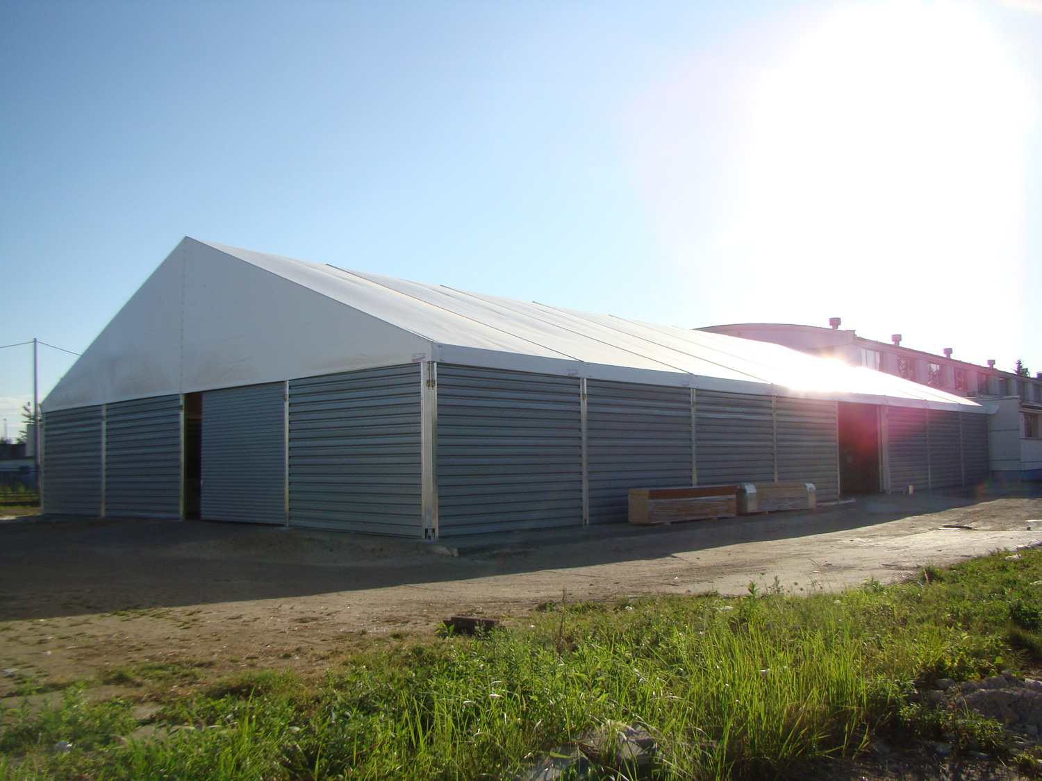 Duża hala namiotowa z szarej blachy trapezowej i białym dachem, widoczna z zewnątrz w słoneczny dzień, częściowo otwarte drzwi przesuwne.