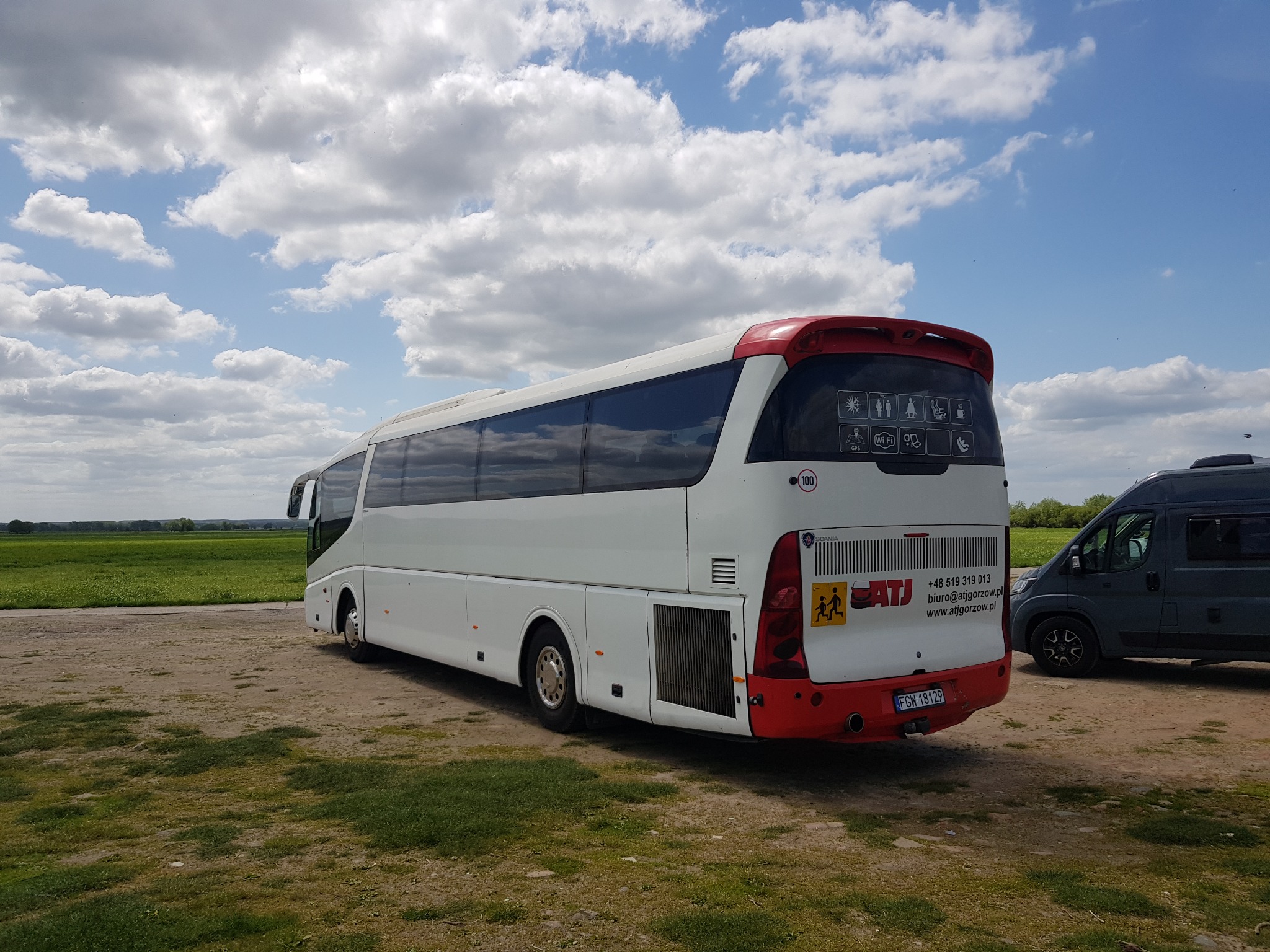 Biały autokar z czerwonym tyłem, zaparkowany na ziemistej powierzchni, w tle zielone pole i błękitne niebo z chmurami. Na tylnej szybie naklejki informacyjne o udogodnieniach dla pasażerów i dane...