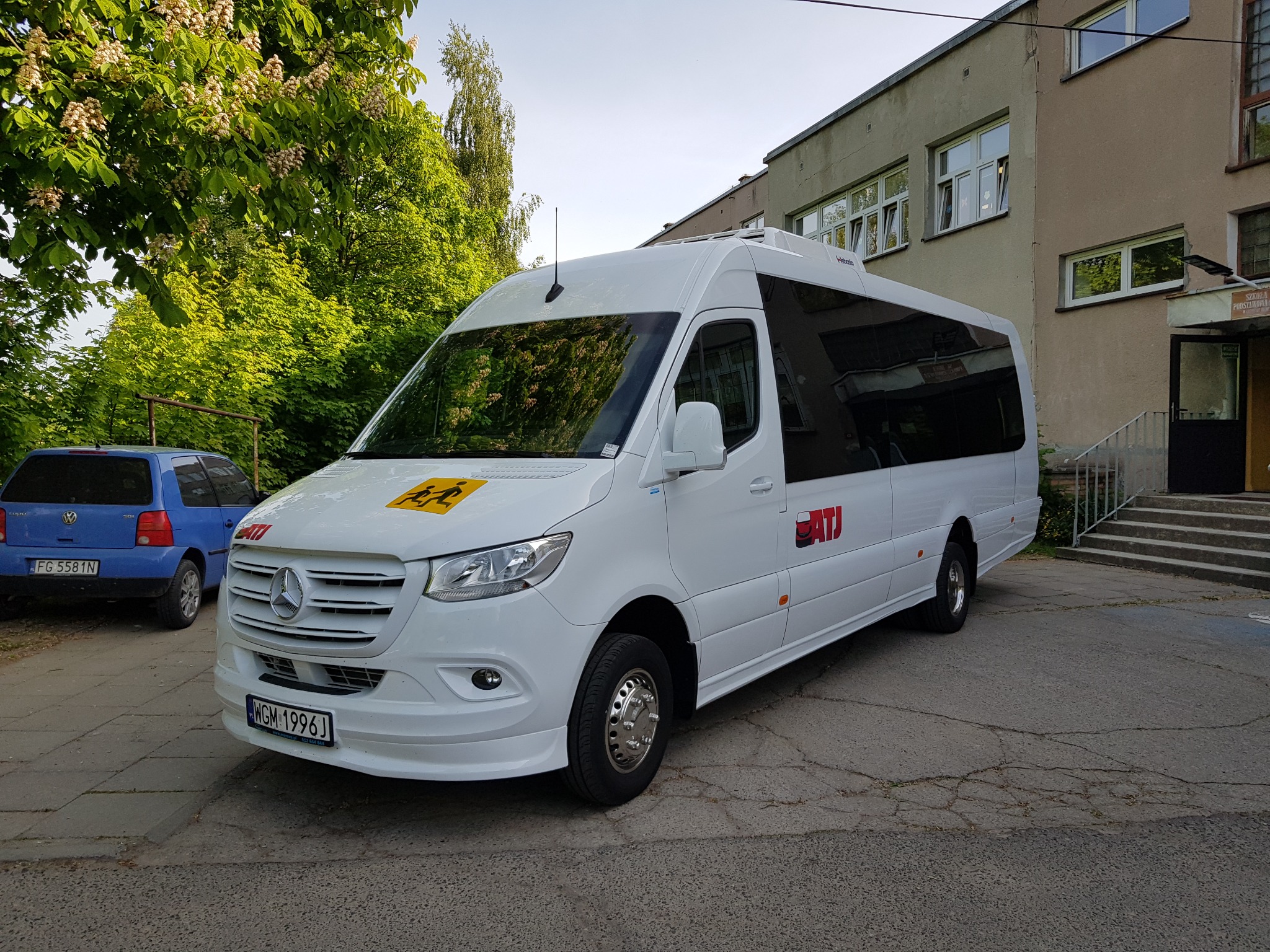 Biały Mercedes Sprinter z naklejką z sylwetkami dzieci na żółtym tle, zaparkowany przed budynkiem szkoły podstawowej, z niebieskim Volkswagenem w tle.