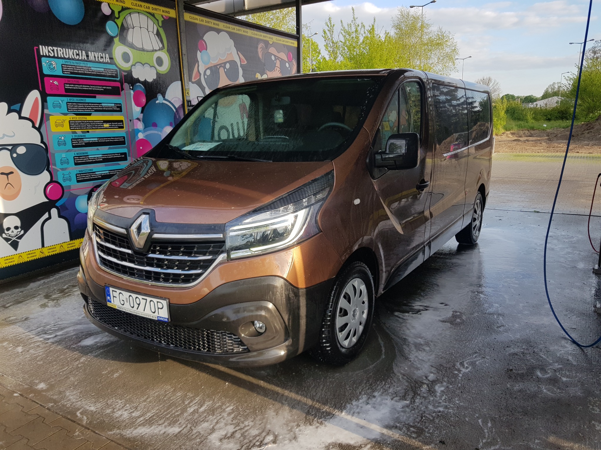 Brązowy bus Renault Trafic myty w samoobsługowej myjni, widoczne resztki piany na karoserii i posadzce, instrukcja mycia na ścianie myjni.