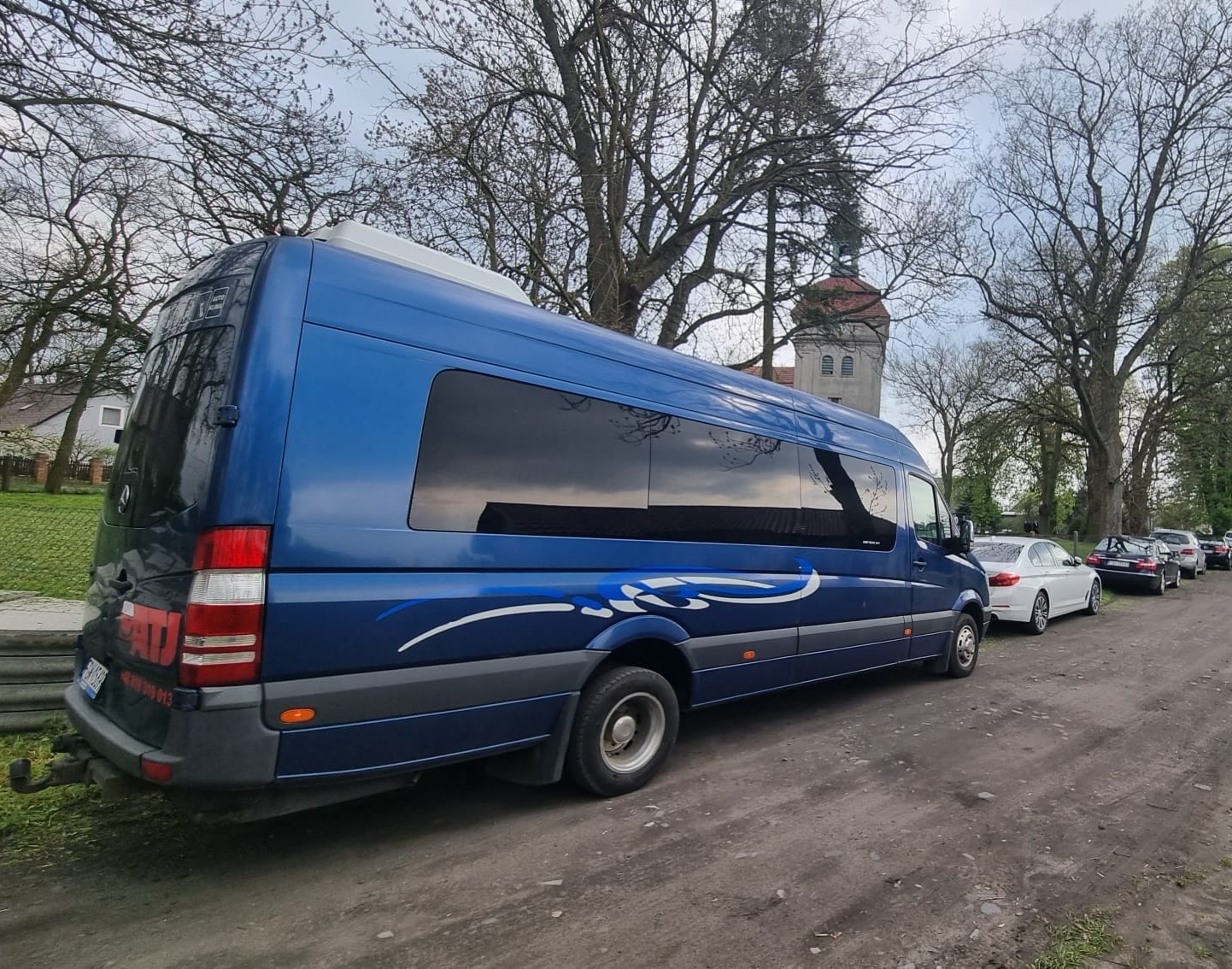 Niebieski bus Mercedes-Benz Sprinter z przyciemnionymi szybami, zaparkowany na żwirowej drodze, zarys kościelnej wieży w tle.