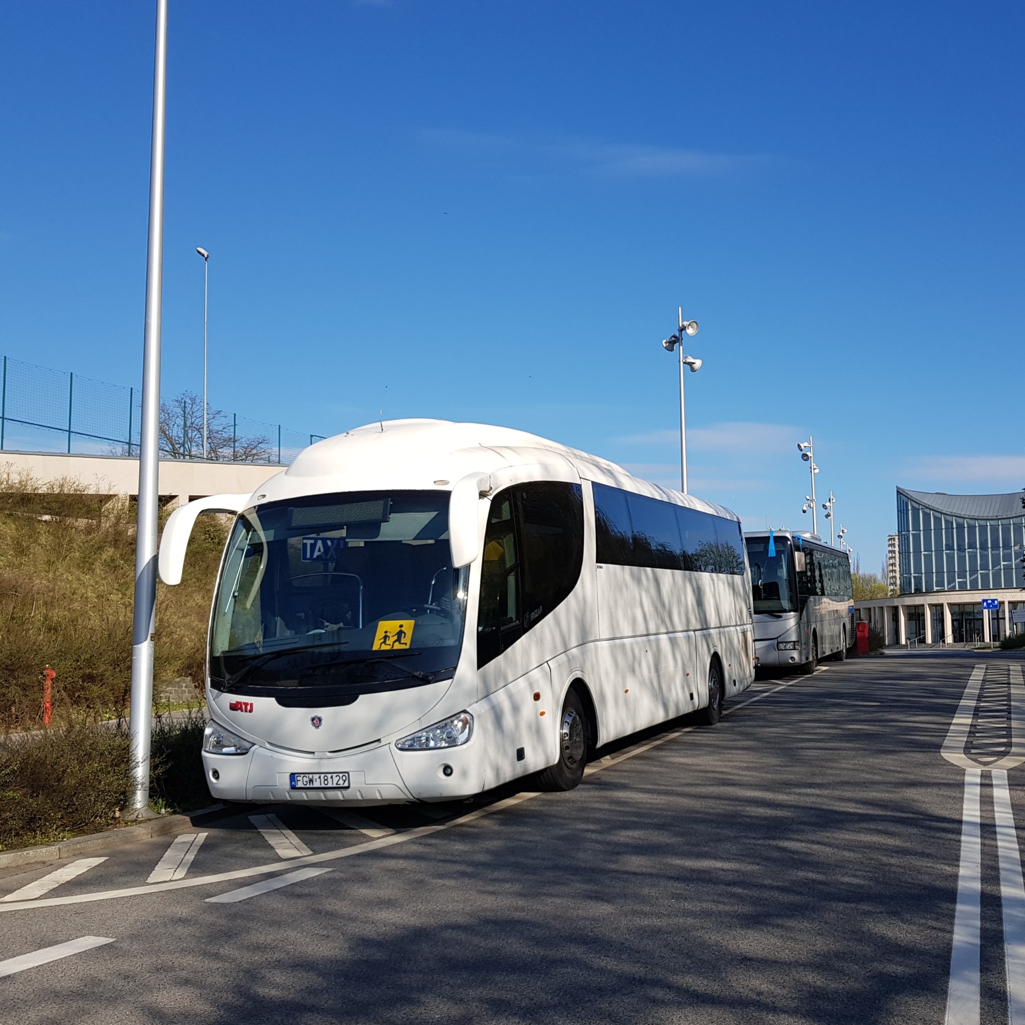 Biały autokar zaparkowany na parkingu, z żółtą naklejką z symbolem dzieci na szybie i tabliczką TAXI w oknie, na tle błękitnego nieba i nowoczesnego budynku.