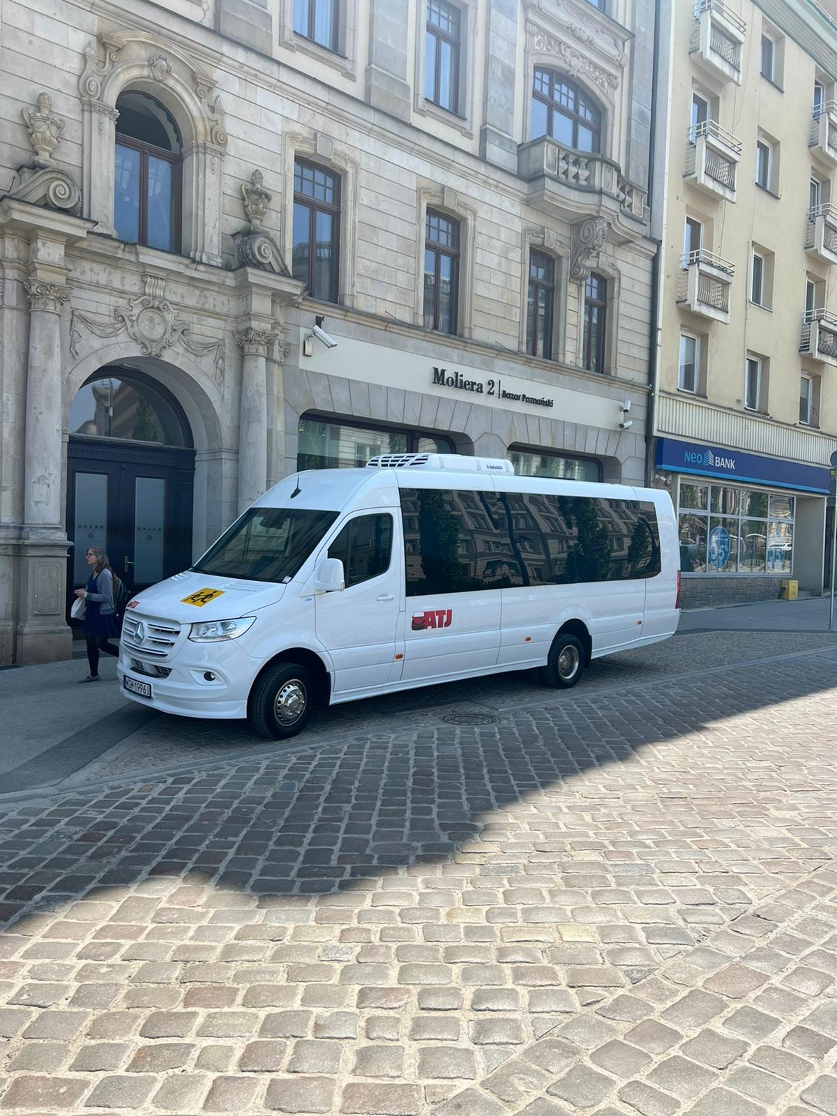 Biały bus Mercedes-Benz z logo firmy ATJ na tle kamienicy z szyldem Moliera 2 i Neo Banku, zaparkowany na brukowanej ulicy.