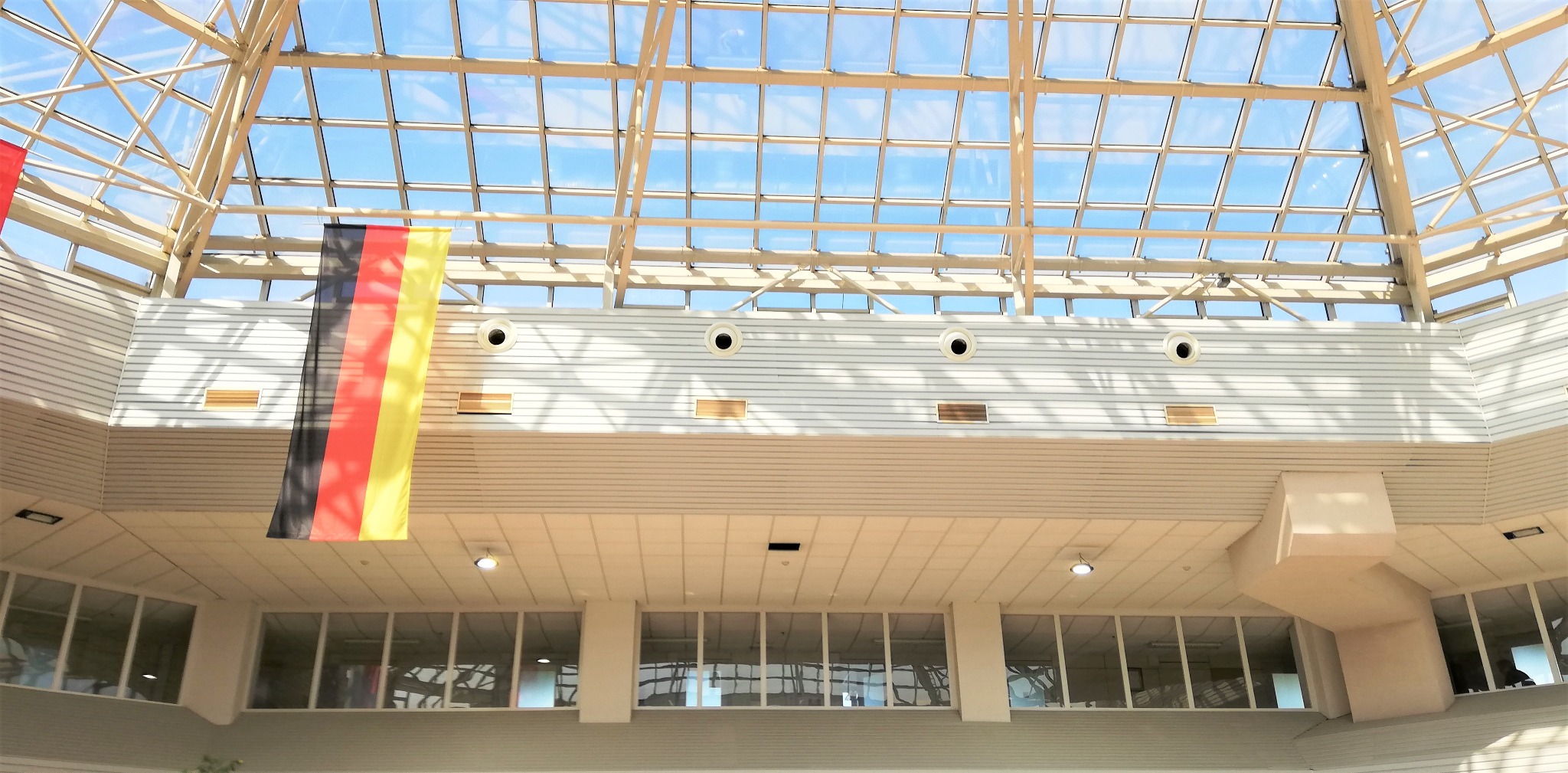 Widok z dołu na przeszklony dach budynku komercyjnego z widocznymi elementami konstrukcyjnymi, oświetleniem i fragmentem flagi Niemiec po lewej stronie.