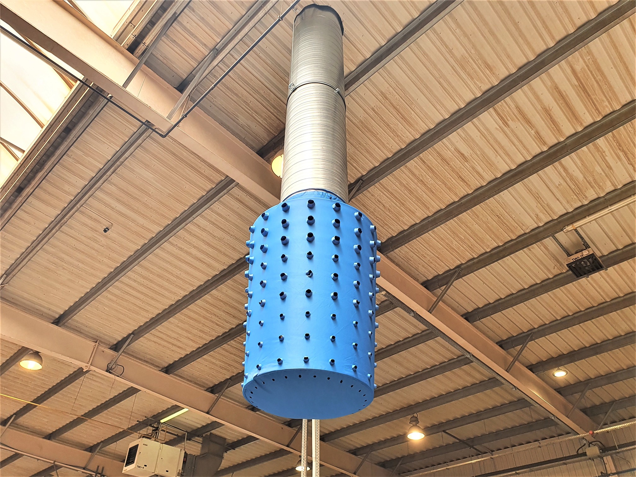 Niebieski cylindryczny element z otworami, część systemu wentylacji przemysłowej, zawieszony pod sufitem hali z widoczną instalacją i oświetleniem.