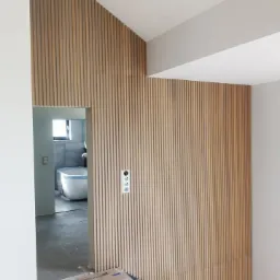 Drewniane lamele pionowe na ścianie, tworzące geometryczny wzór, z widocznym fragmentem łazienki w tle i zabezpieczonymi schodami na pierwszym planie.