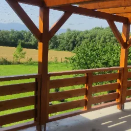 Drewniany taras z widokiem na zielone pola i drzewa, konstrukcja z belek i balustrad w ciepłym odcieniu brązu, słoneczny dzień.