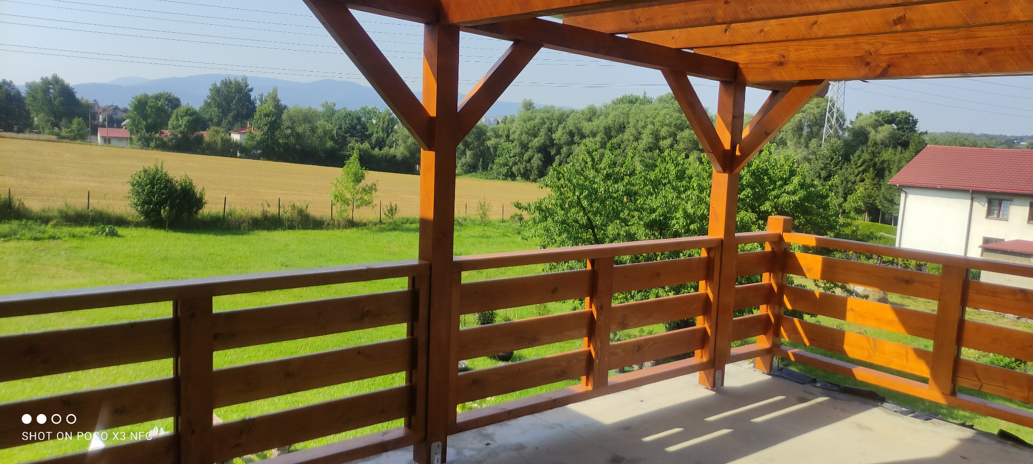 Drewniany taras z widokiem na zielone pola i drzewa, konstrukcja z belek i balustrad w ciepłym odcieniu brązu, słoneczny dzień.