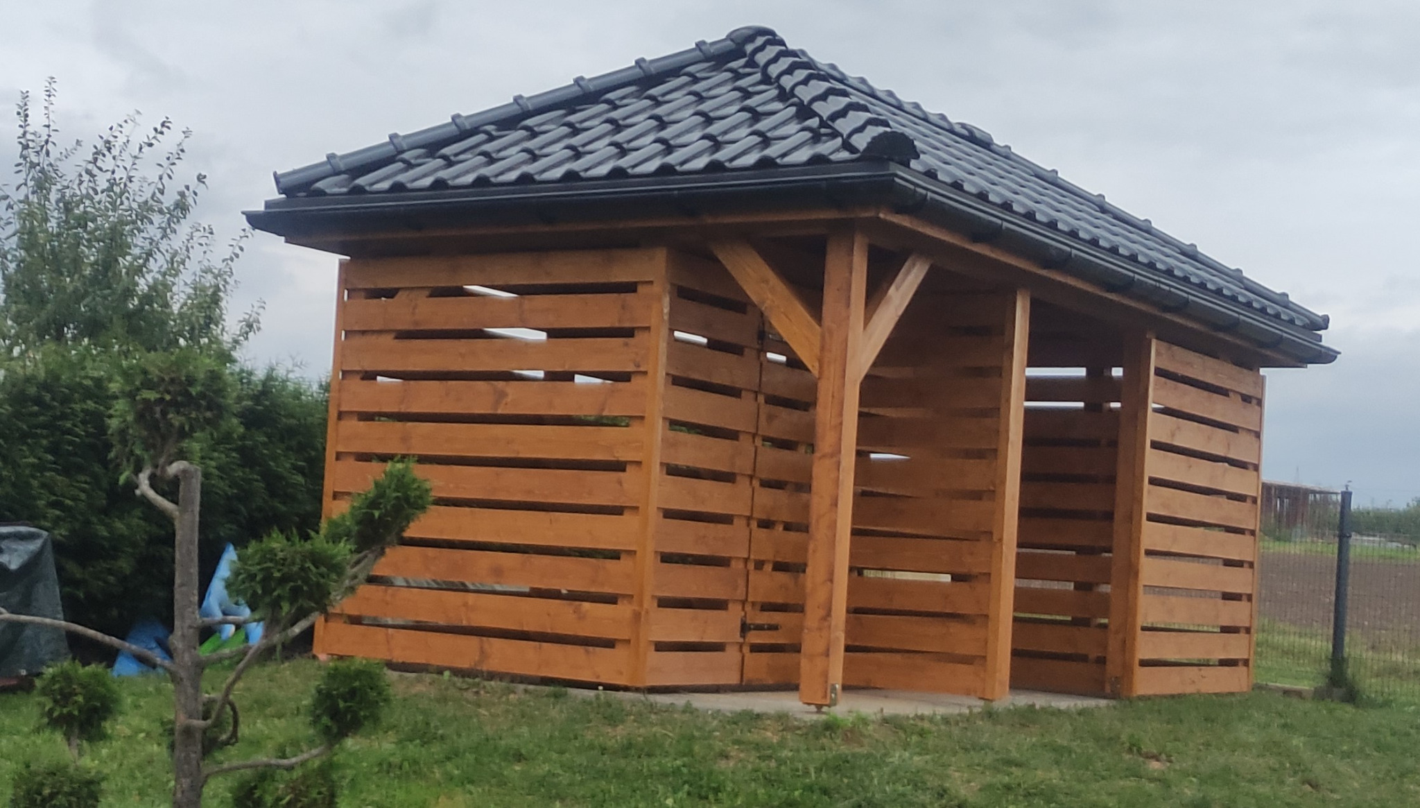 Drewniana altana z ażurowymi ścianami i czarnym dachem z dachówki, widoczna z zewnątrz na tle zielonego trawnika i pochmurnego nieba.