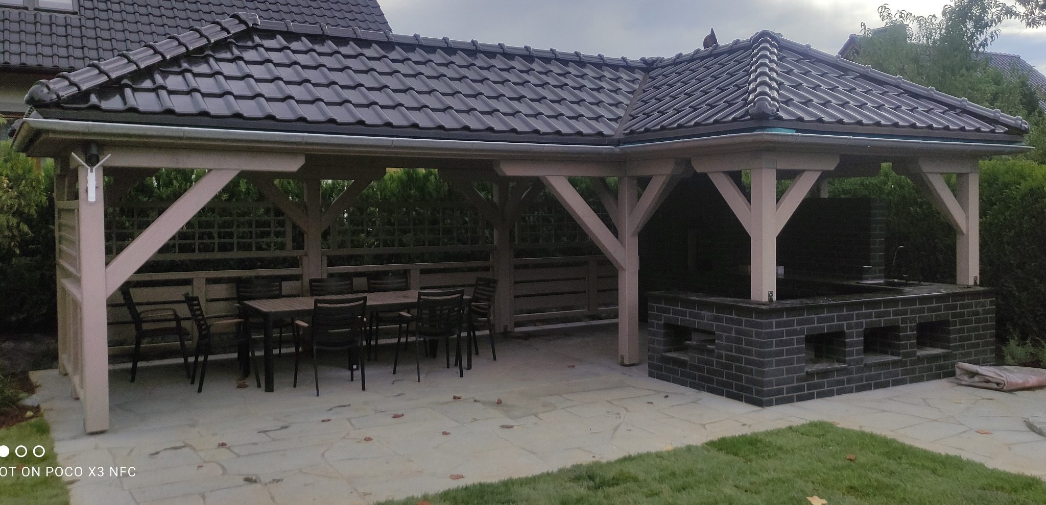 Zadaszona konstrukcja z ciemną dachówką, zawierająca stół z krzesłami oraz murowany grill z ciemnej cegły, umieszczona na kamiennej posadzce w rogu działki.