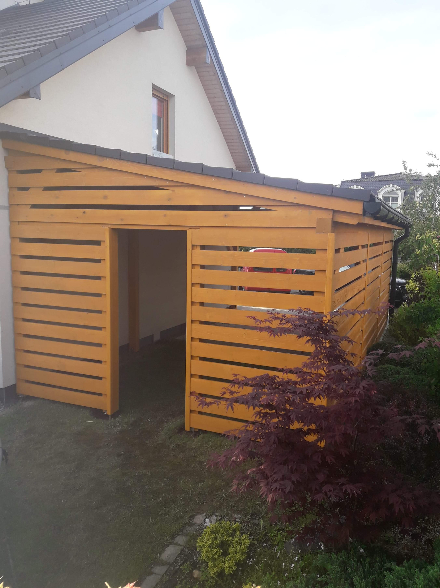 Drewniana konstrukcja z poziomych desek, przylegająca do budynku, z otwartym wejściem i widocznym wnętrzem, na tle zielonego trawnika i krzewów.