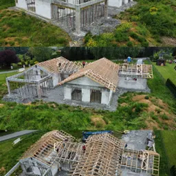 Widok z drona na trzy domy murowane w różnym stadium budowy: jeden w stanie surowym otwartym z widocznymi fundamentami i ścianami z bloczków, drugi z konstrukcją dachu w trakcie montażu, a trzeci...