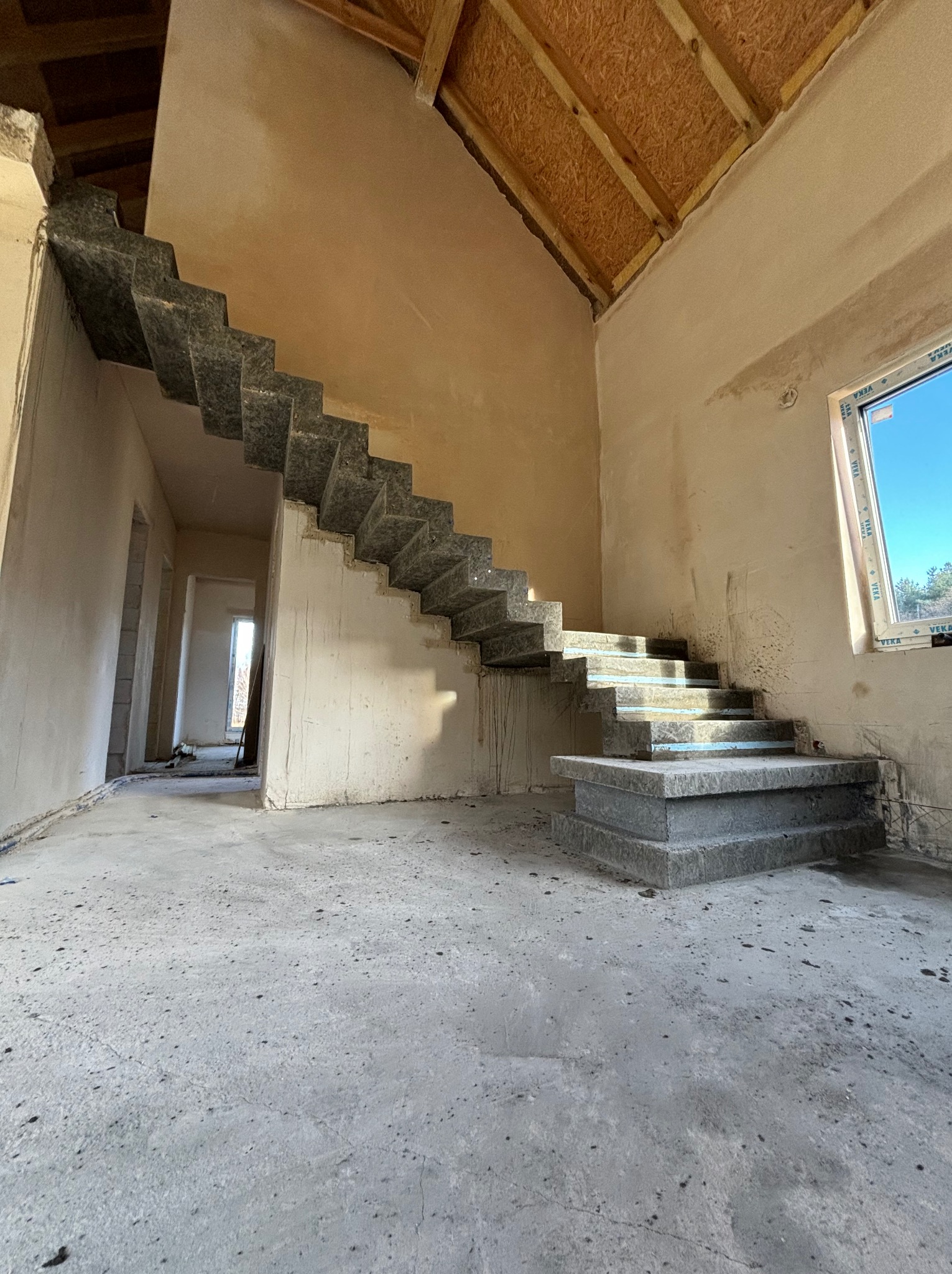 Surowe, niedokończone schody w nowym domu, widoczne szare stopnie bez balustrady, surowe ściany w odcieniach beżu, drewniane elementy dachu i okno z widokiem na zieleń na zewnątrz.